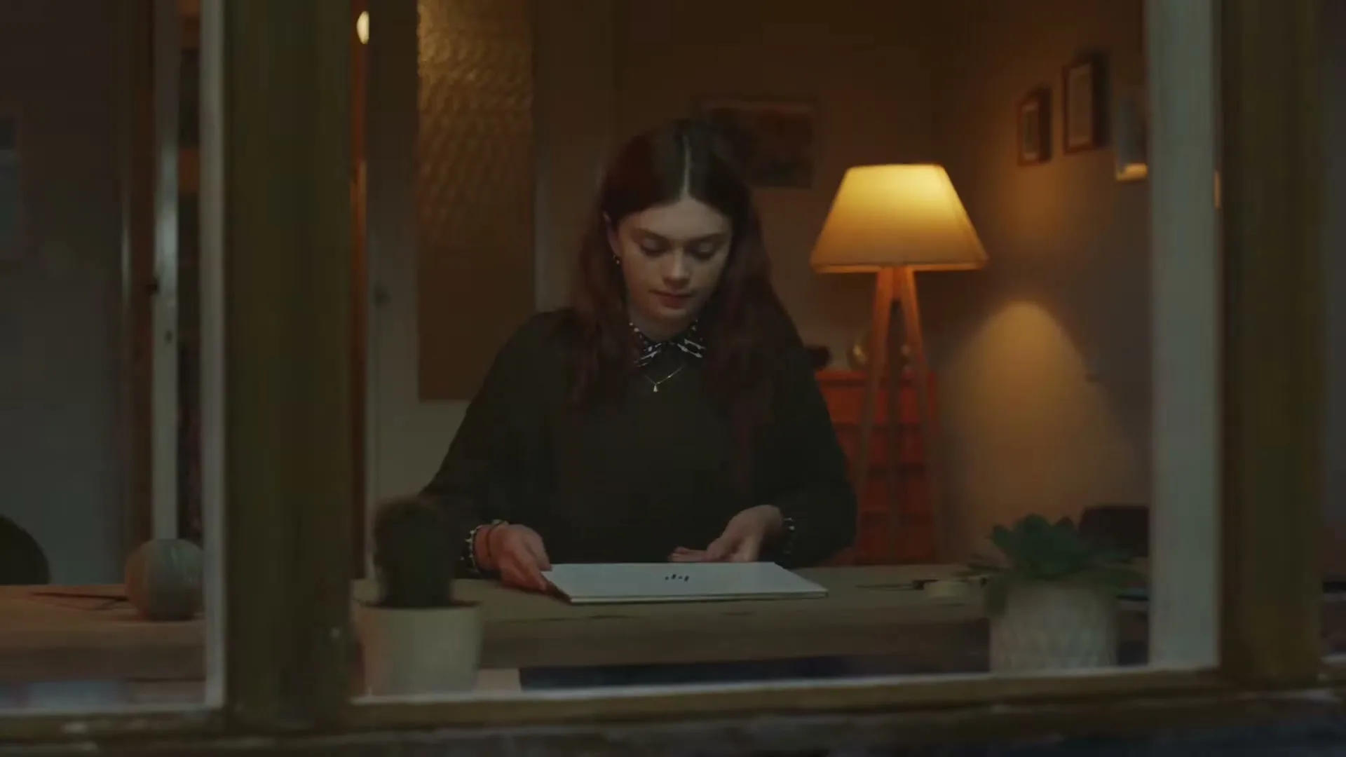 A young woman with long brown hair sits at a table, opening a silver laptop, viewed through a window at night.