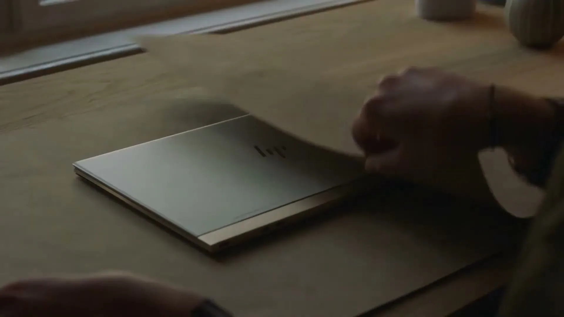 A person's hands carefully peel back brown wrapping paper from a silver HP laptop on a wooden table next to a window.