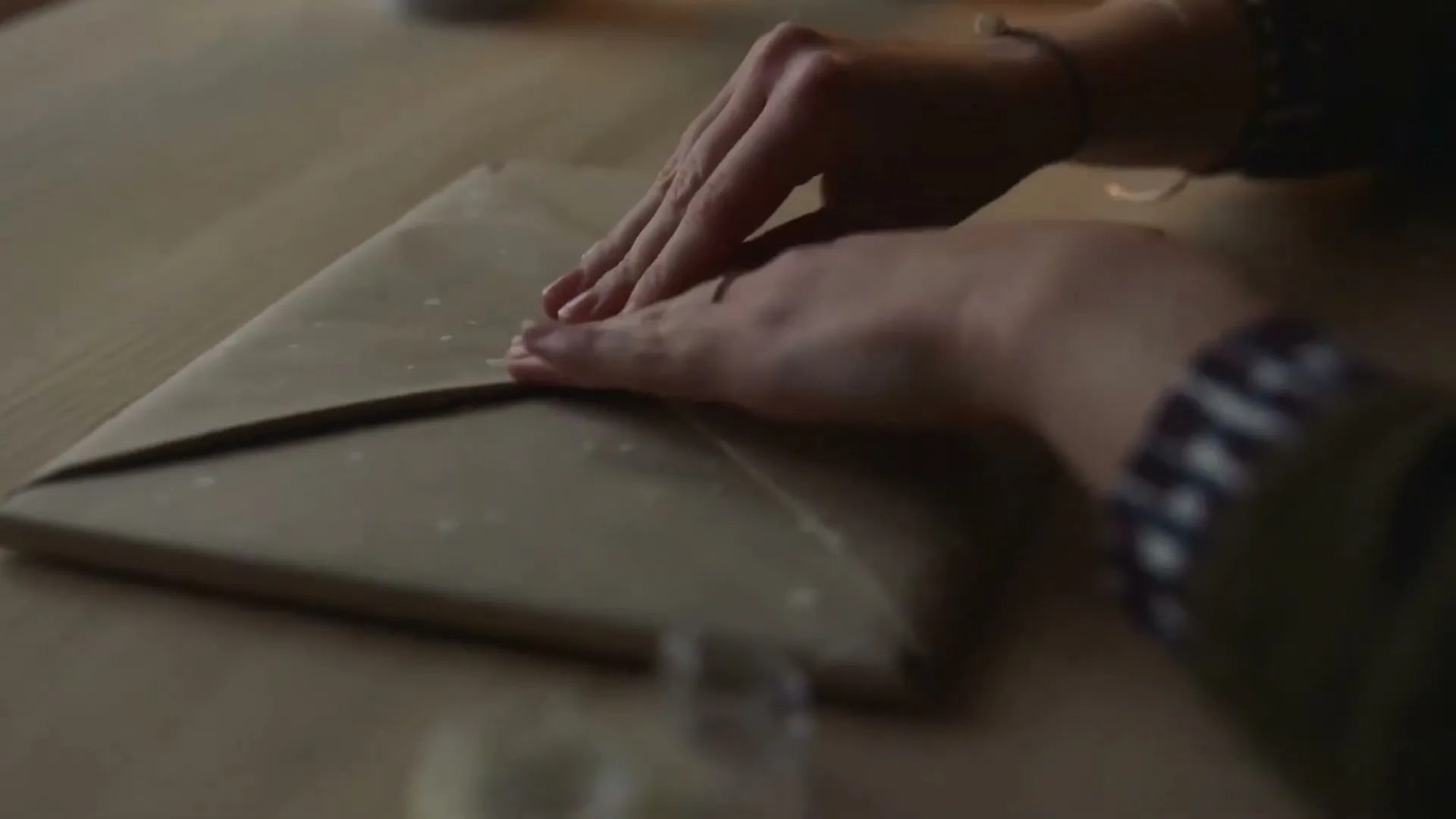 A person's hands are seen wrapping a gift in brown paper with white dots on a wooden table.
