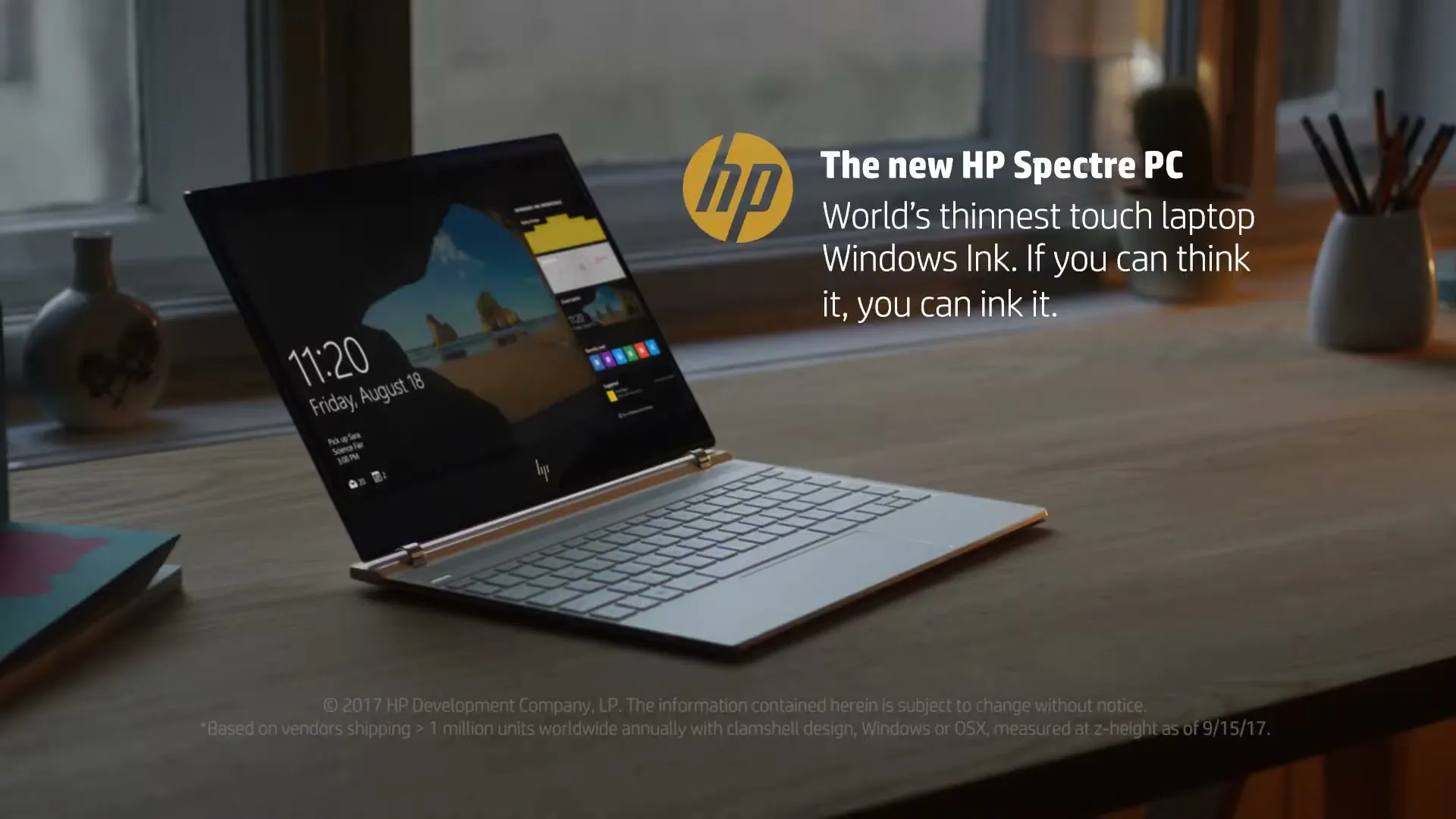 A sleek, open HP Spectre PC laptop sits on a wooden desk with a window in the background, displaying the time and date on its screen.