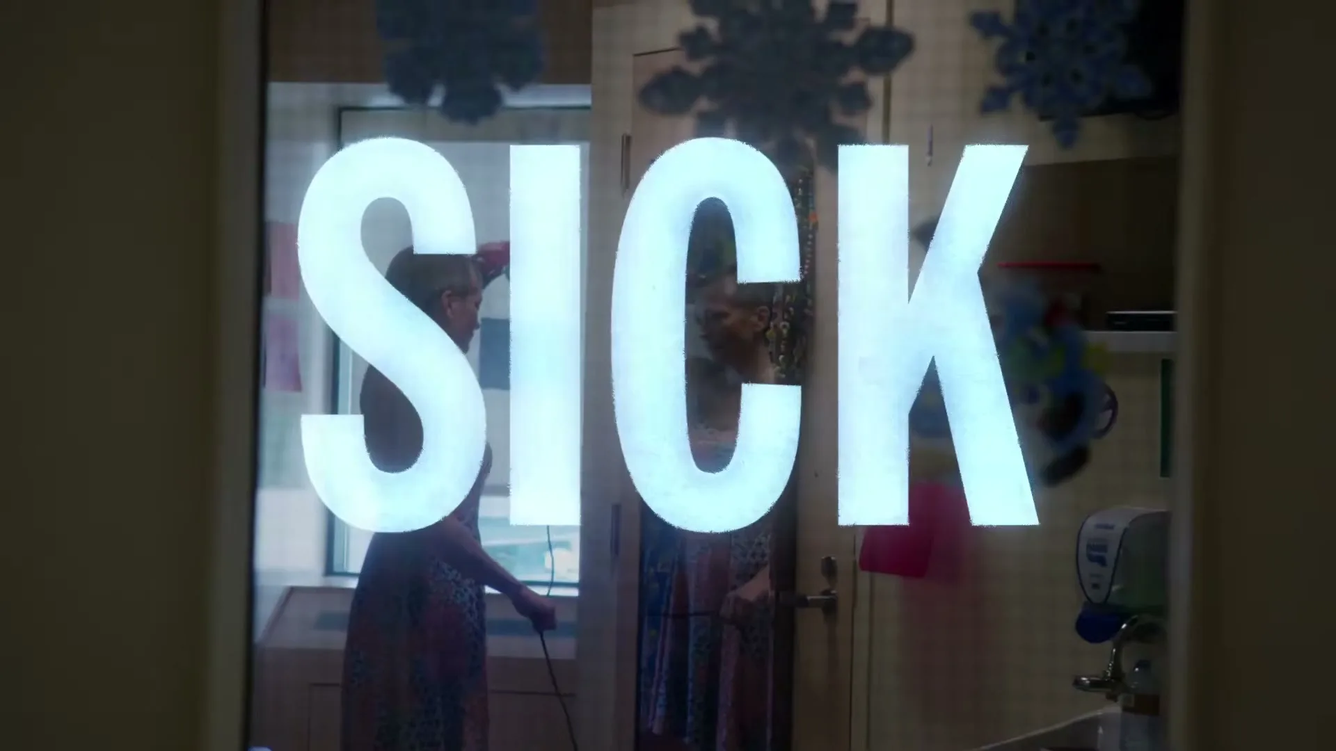 The word "SICK" is displayed prominently on a glass door, with a blurry reflection of a person visible behind it in a hospital room.