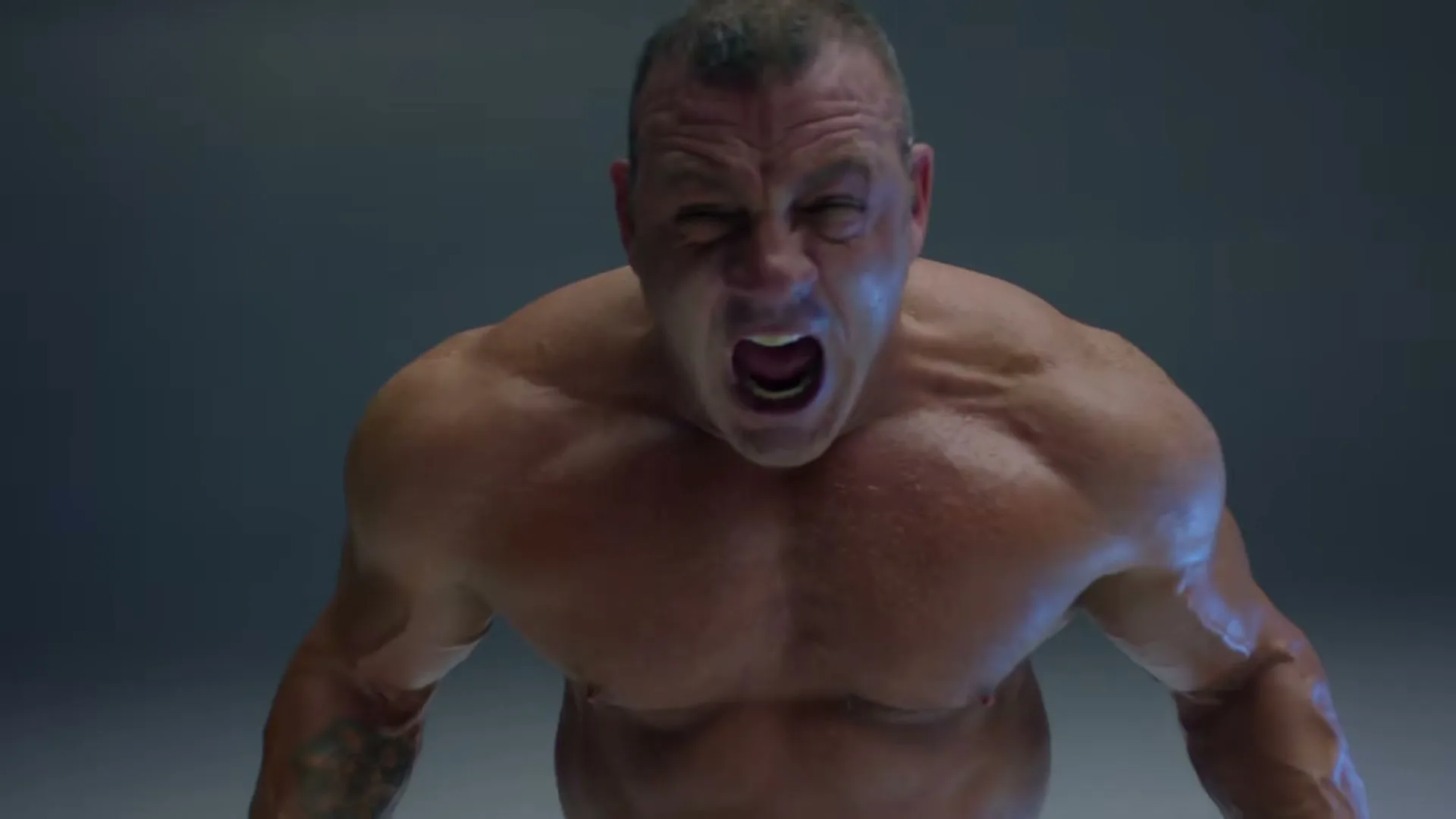 A muscular, shirtless man with a mohawk screams intensely at the camera in a studio setting, his body glistening with sweat.