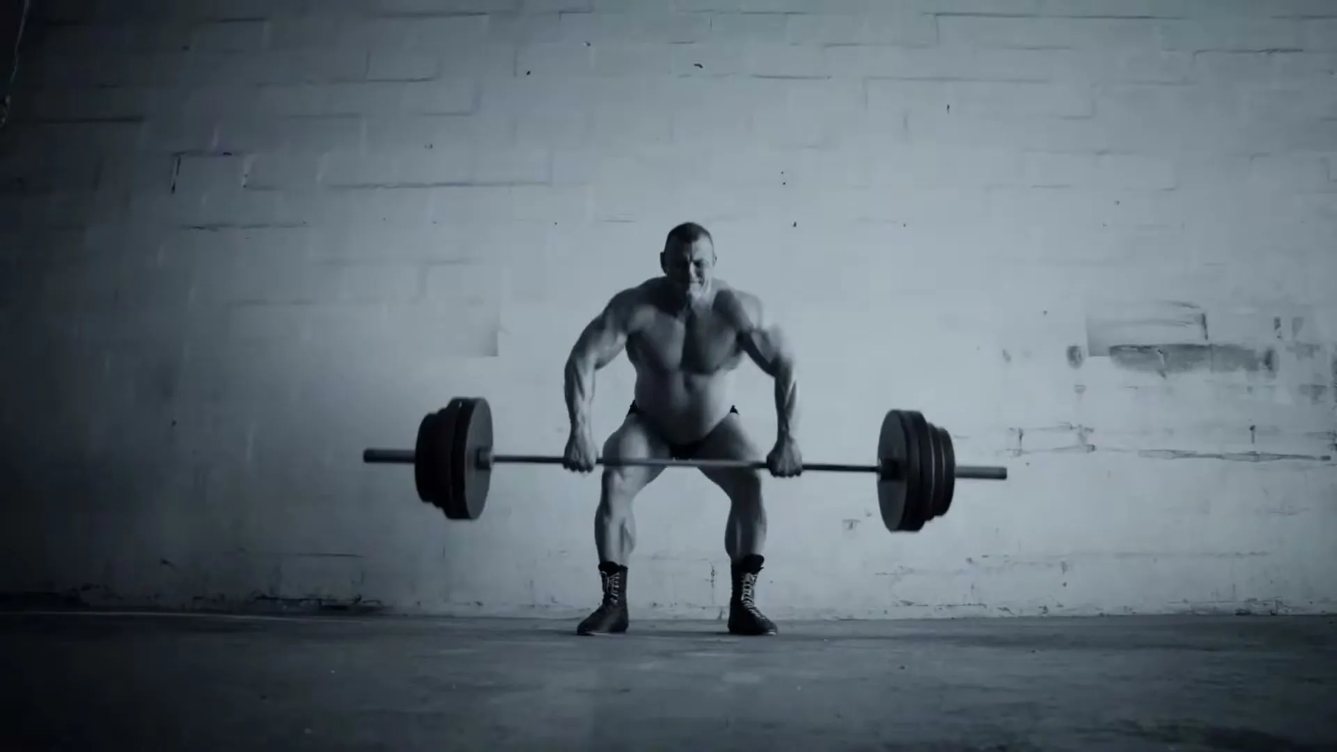 A muscular male bodybuilder in black briefs and boots lifts a heavy barbell in a stark, industrial-looking room.