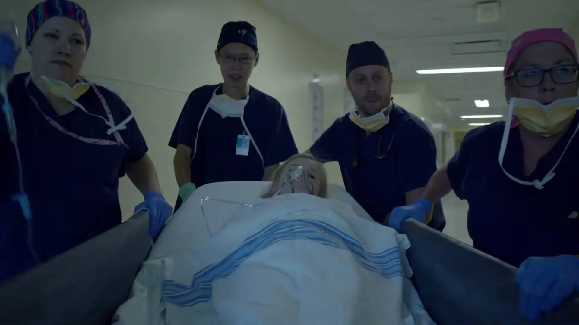 Four medical professionals in scrubs rush a young patient on a gurney down a hospital hallway, looking determined and focused.