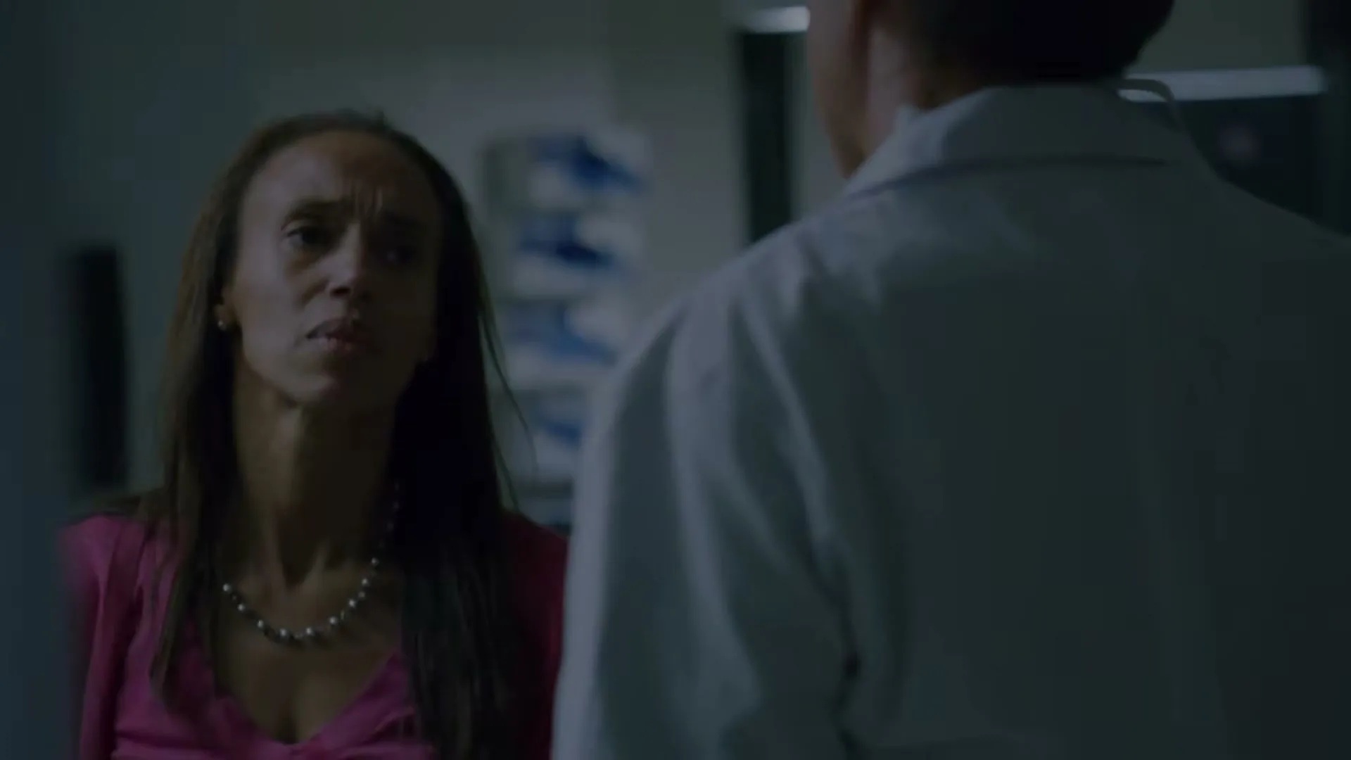 A woman with long dark hair and a pink top looks concerned while talking to a person in a white lab coat in a hospital hallway.