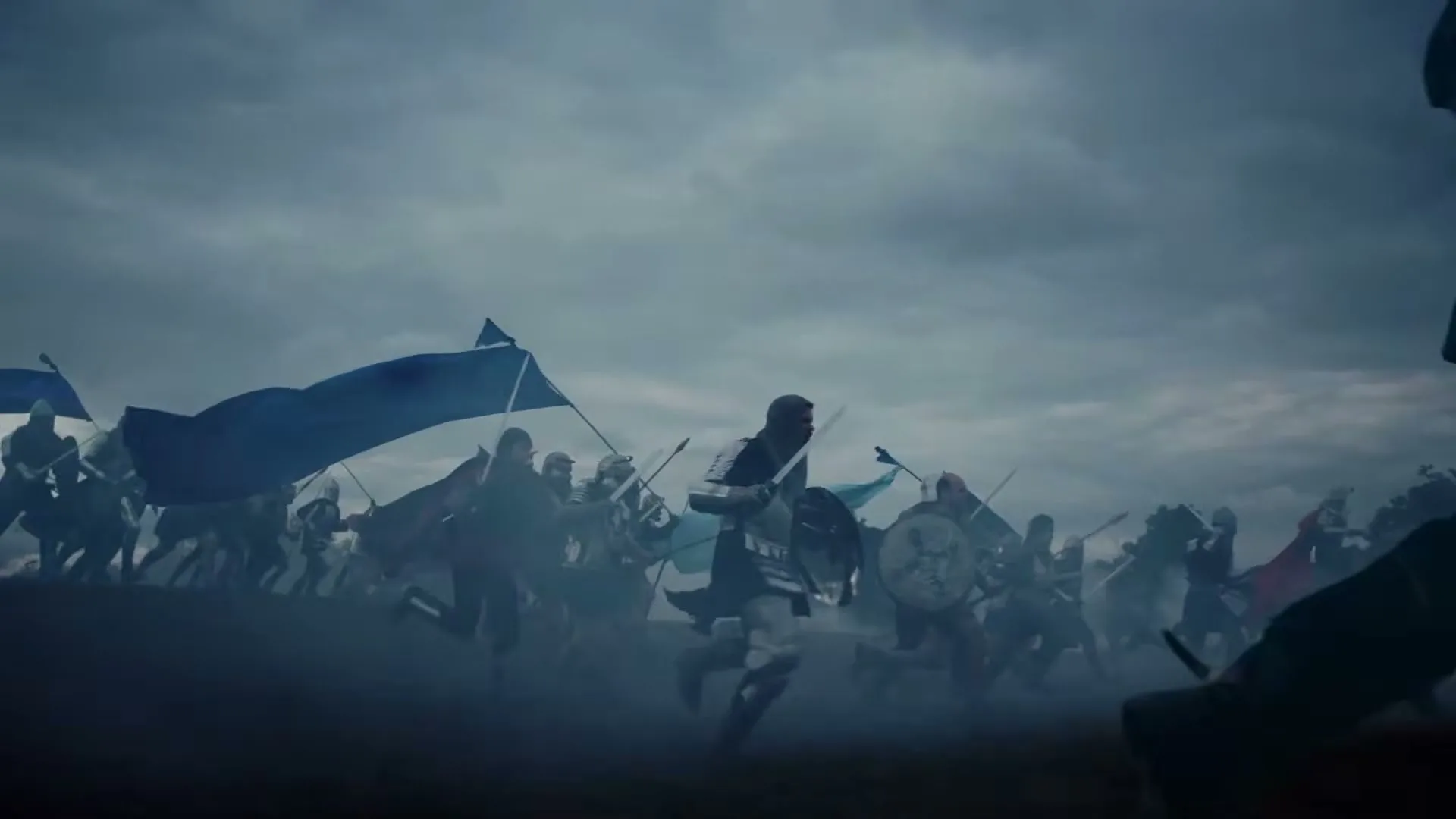 A large army of medieval warriors, carrying blue flags and shields, charges across a smoky battlefield under a cloudy sky.