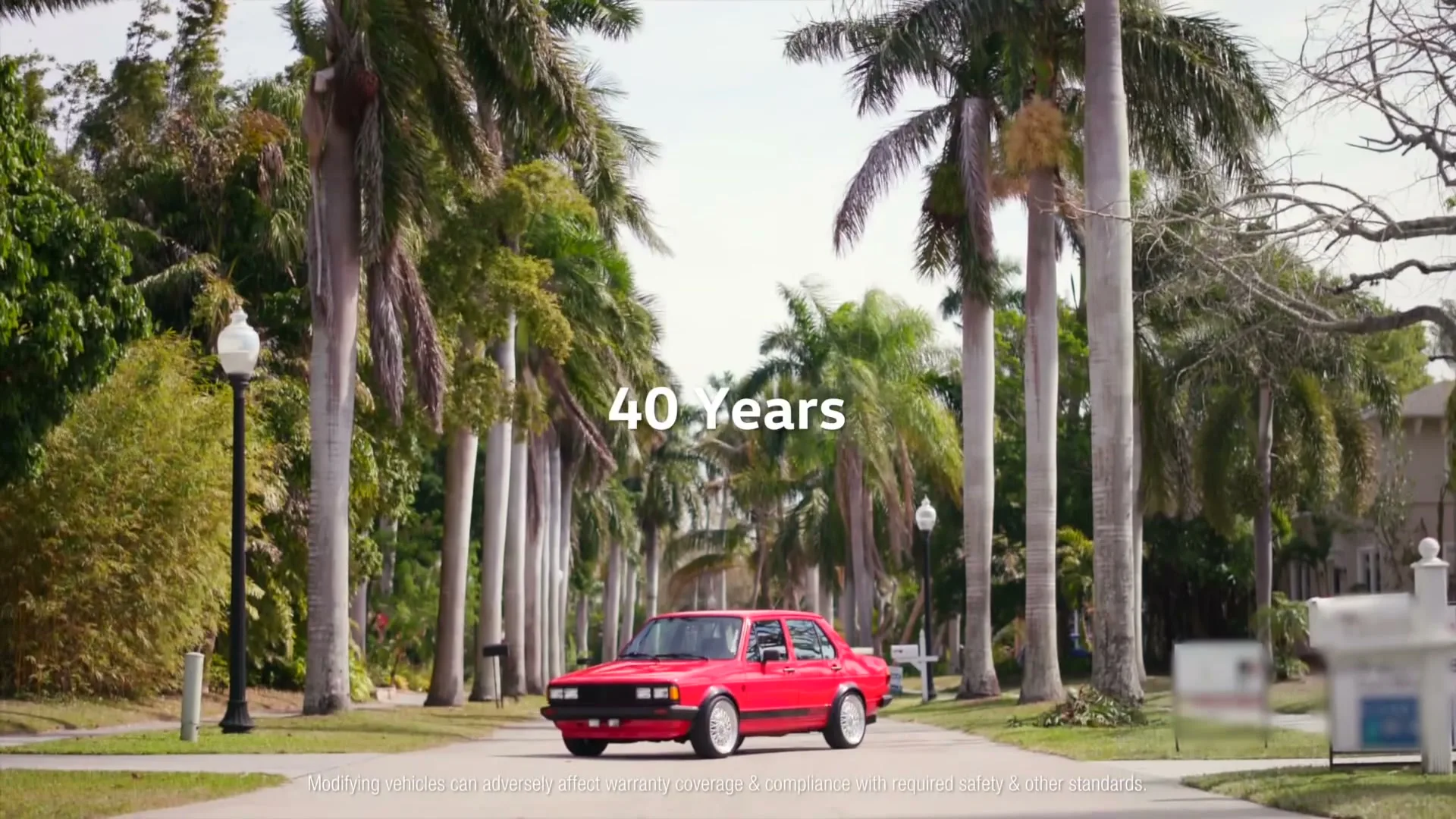A red Volkswagen car drives slowly down a palm tree-lined suburban street on a bright, sunny day.