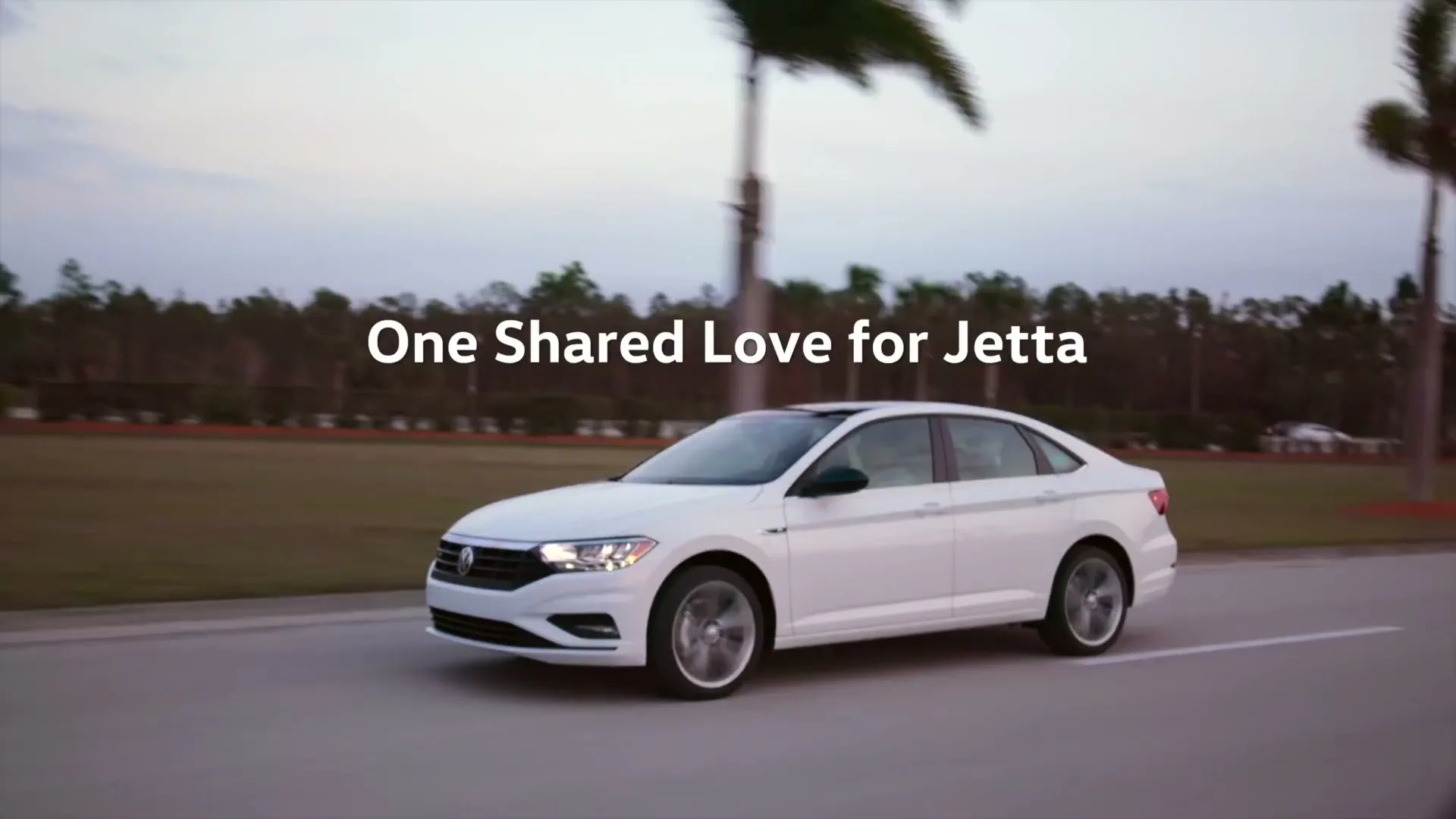 A white Volkswagen Jetta drives on a road with palm trees in the background during golden hour.
