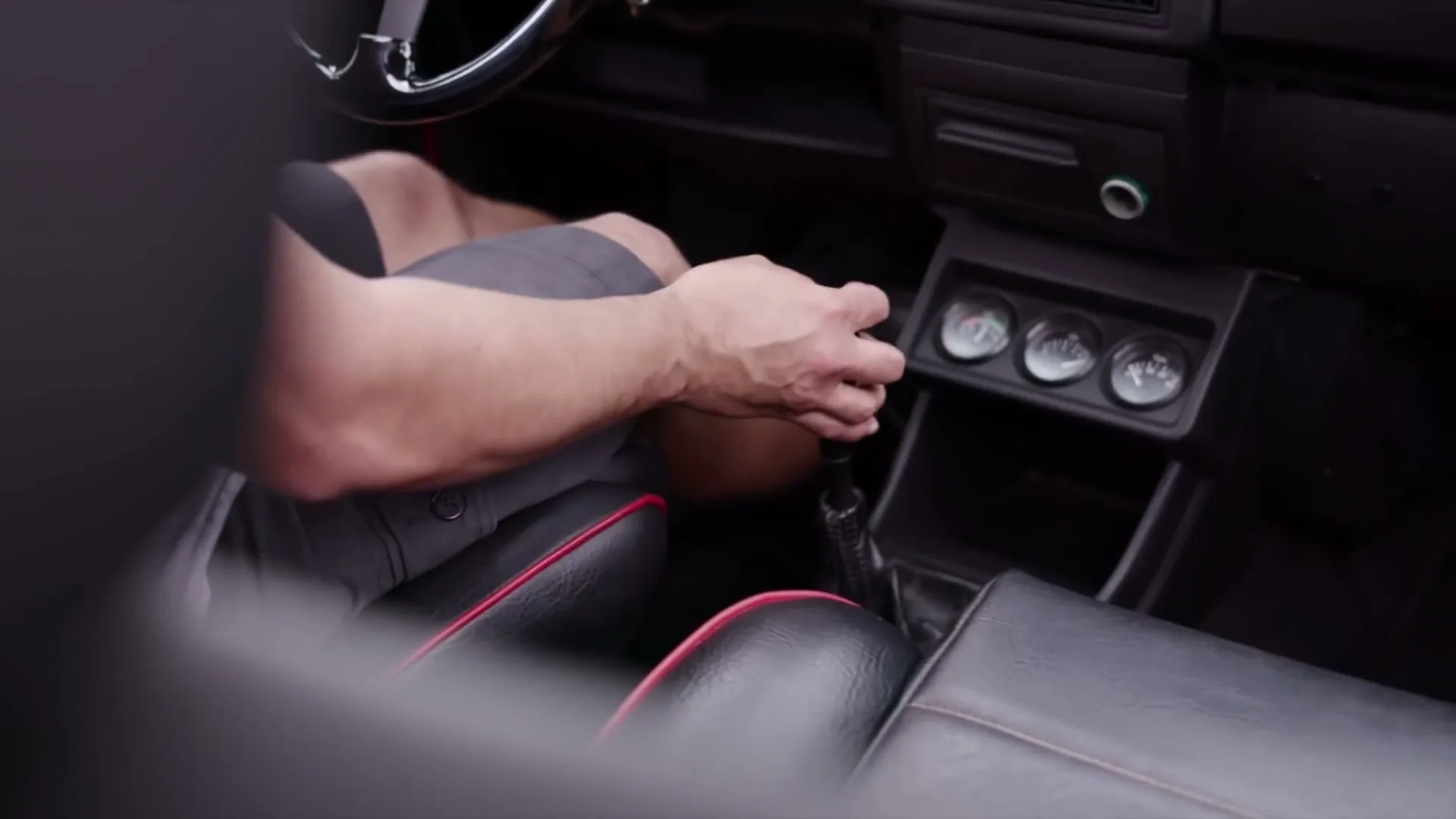 A man's arm and hand are seen shifting gears in a car, with a glimpse of his legs and the car's interior.