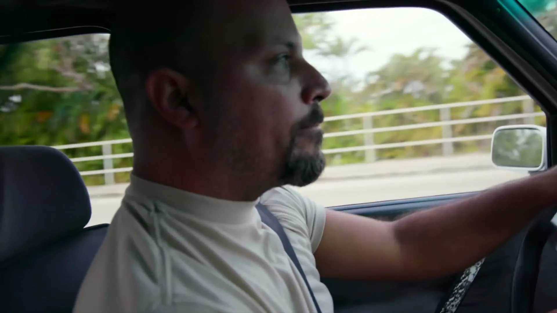 A bald man with a beard drives a car, looking forward with a serious expression, on a sunny day.