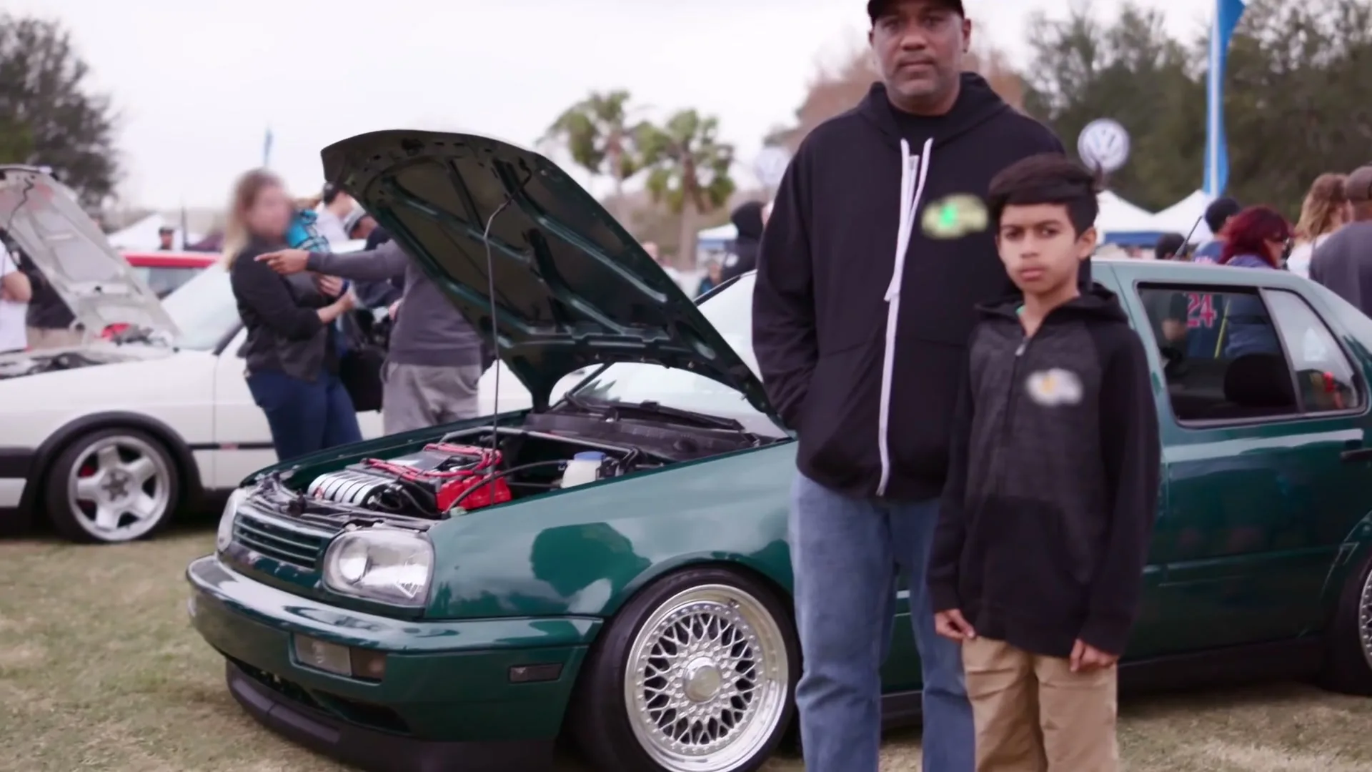A father and son stand next to a dark green Volkswagen car with its hood open at an outdoor car show during the day.