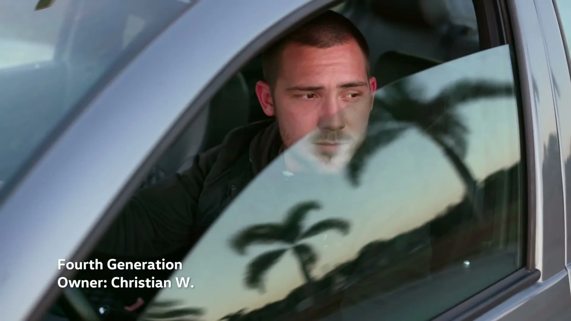 A young man with short hair and a beard is seen through a car window, looking forward, then turning to smile at the camera, with palm trees reflected in the glass.