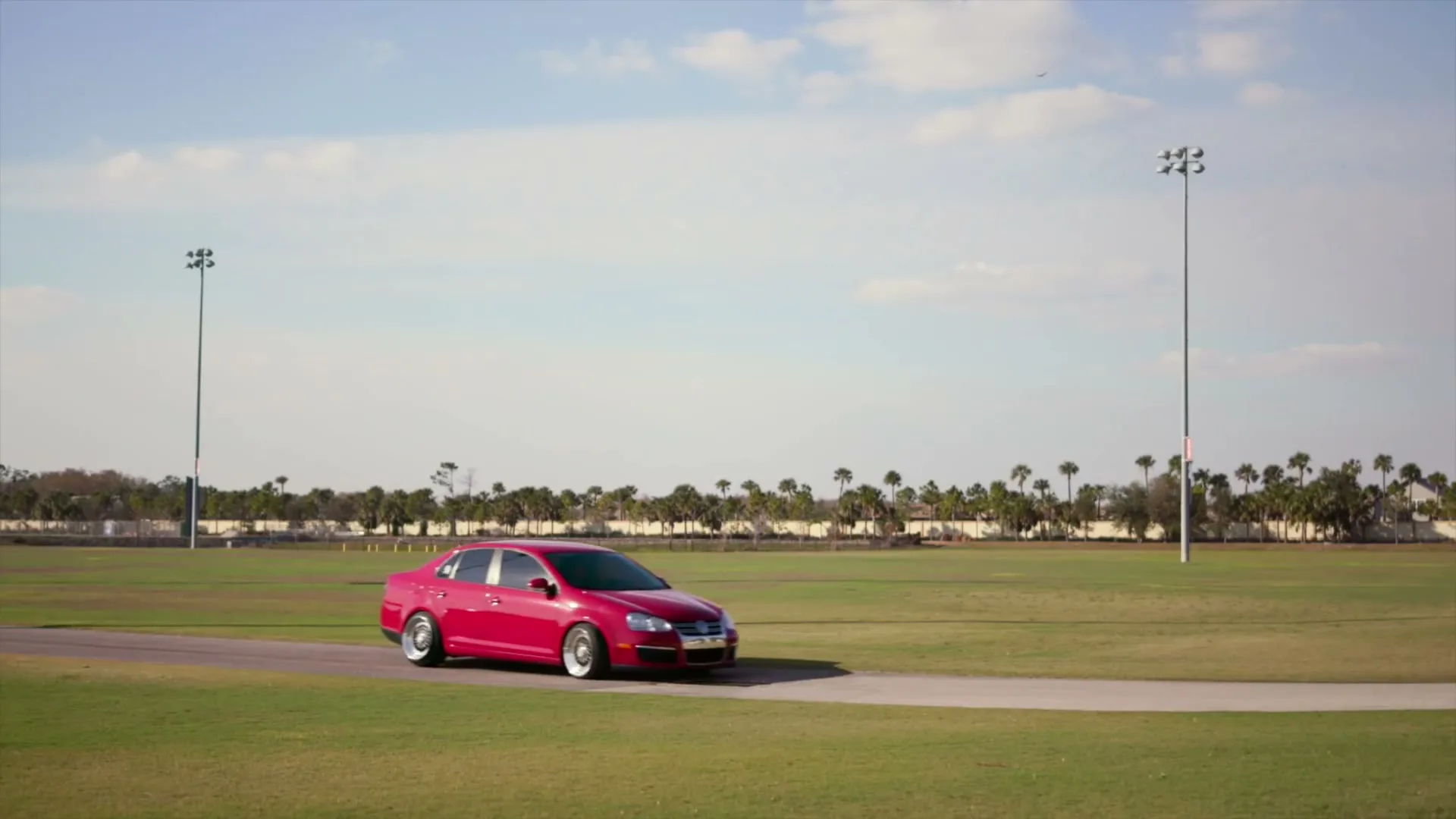 A red Volkswagen Jetta is parked on a paved path in a large grassy field under a bright blue sky with scattered clouds.