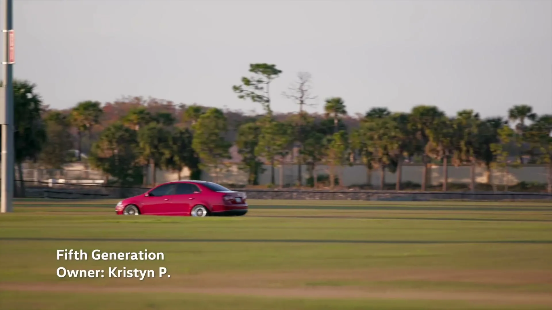 A red Volkswagen Jetta drives across a wide, flat green field with palm trees and a fence in the background under a clear sky.