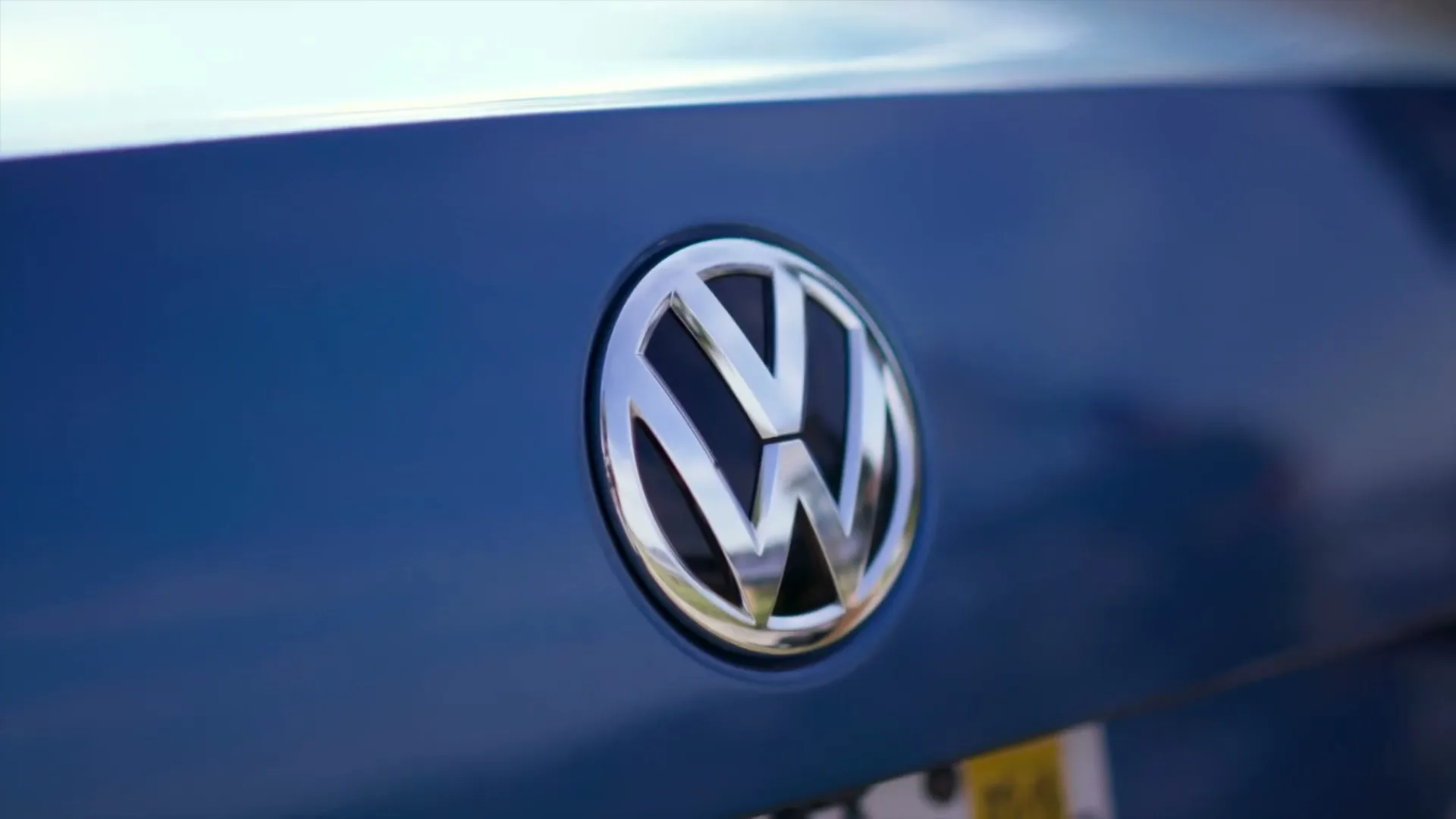 A close-up shot of the shiny silver Volkswagen logo on the blue trunk of a car, with a blurred background of the sky and a license plate.