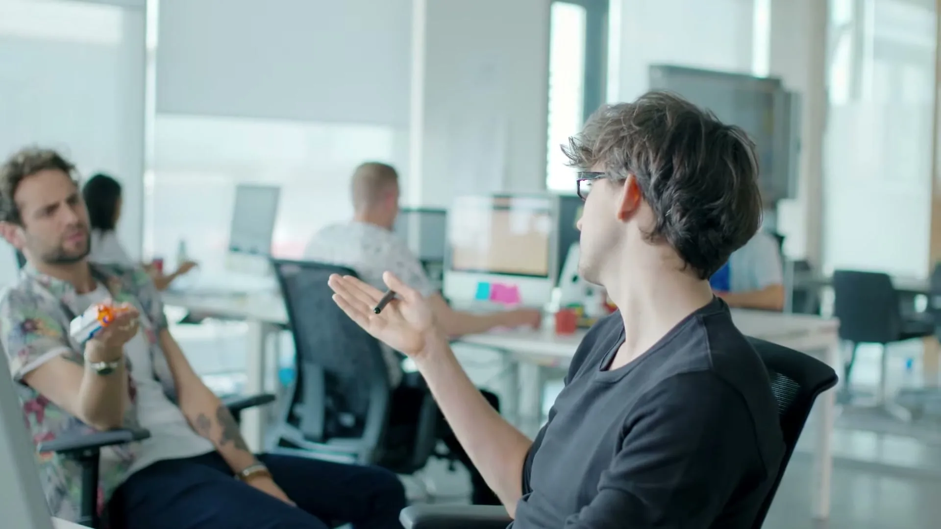Two men in an office, one with curly hair and glasses, the other with a beard and a Hawaiian shirt, are talking to each other while other people work in the background.