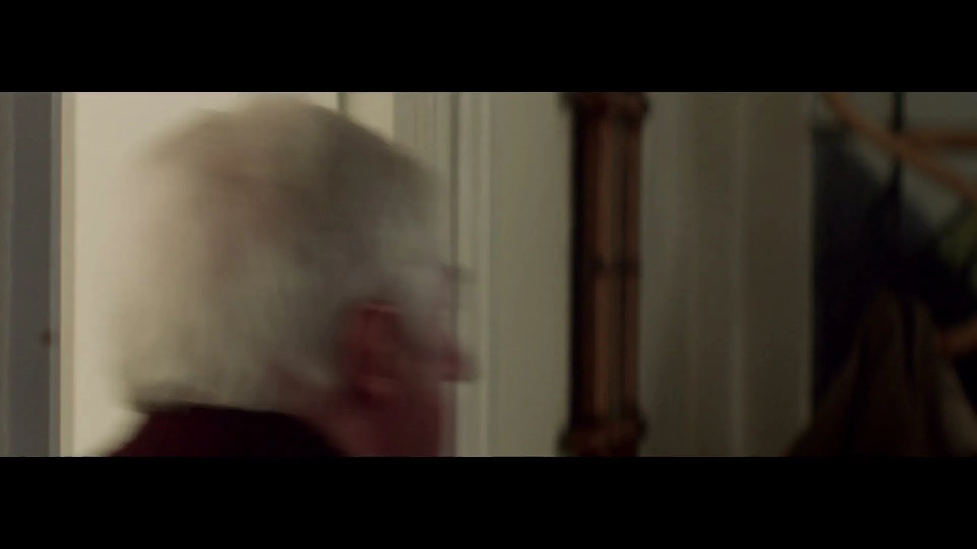 A man with white hair and a maroon sweater walks past a white door and a wooden coat rack in a home.