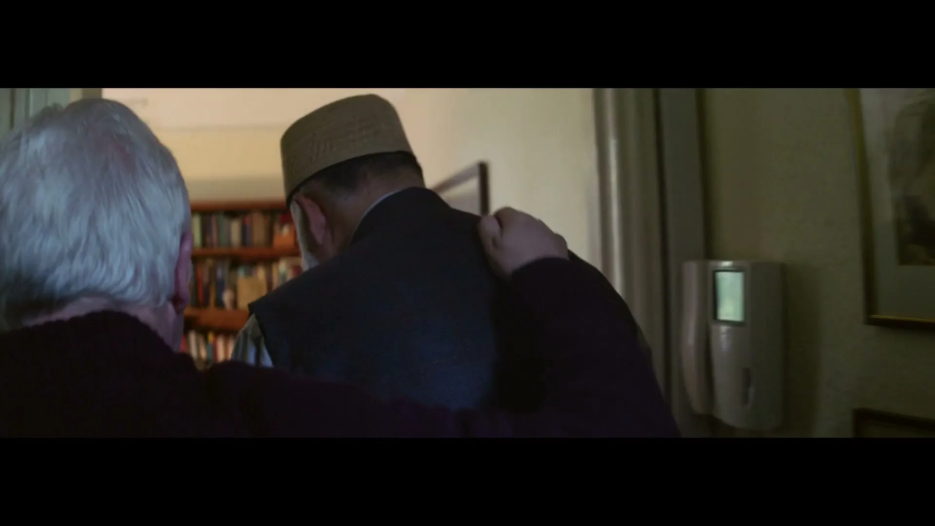 An older person with white hair puts their arm around another older person wearing a cap, walking into a room with bookshelves.
