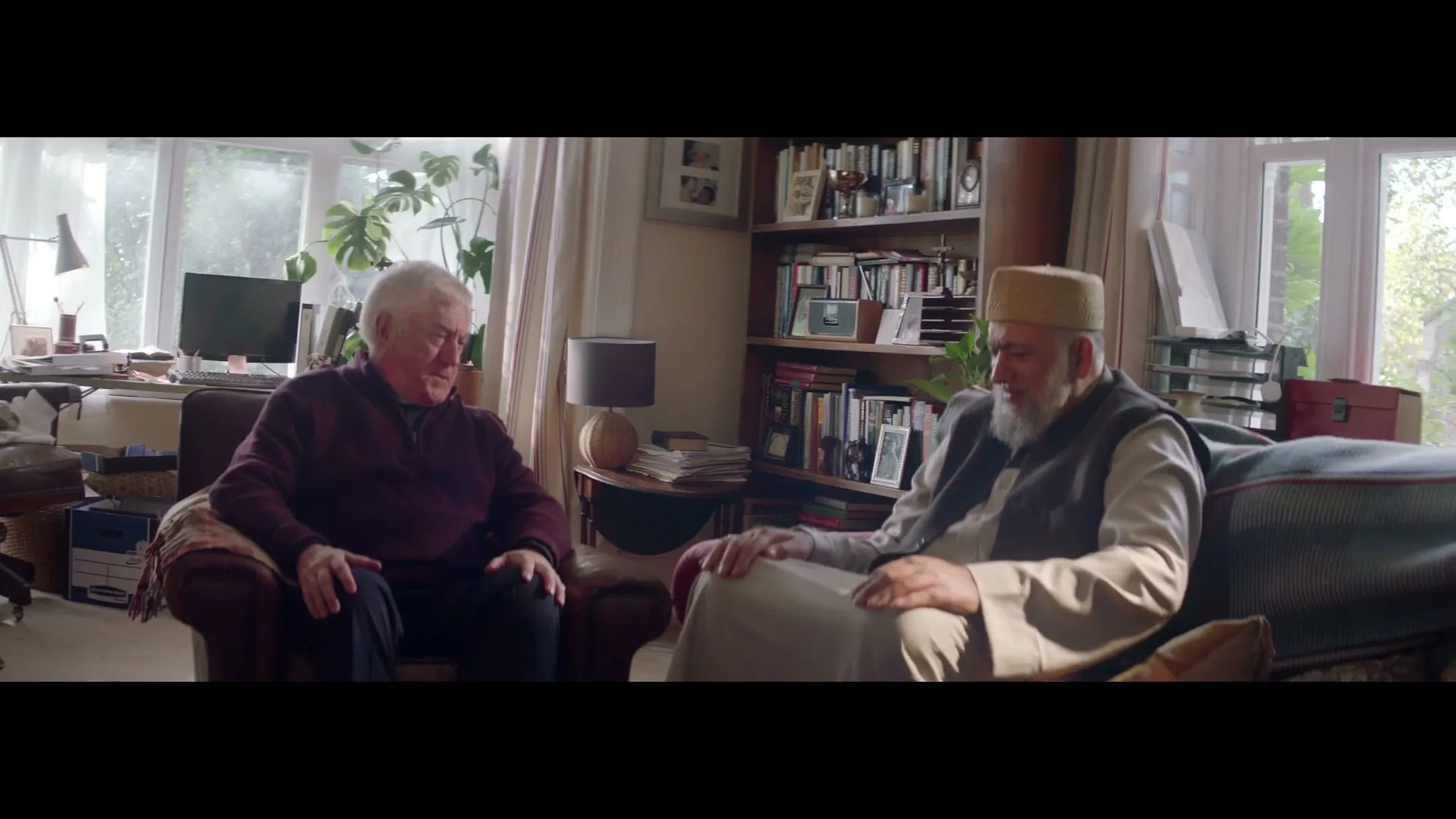 Two elderly men, one in a maroon sweater and the other in traditional attire, sit in a cozy living room, conversing warmly.