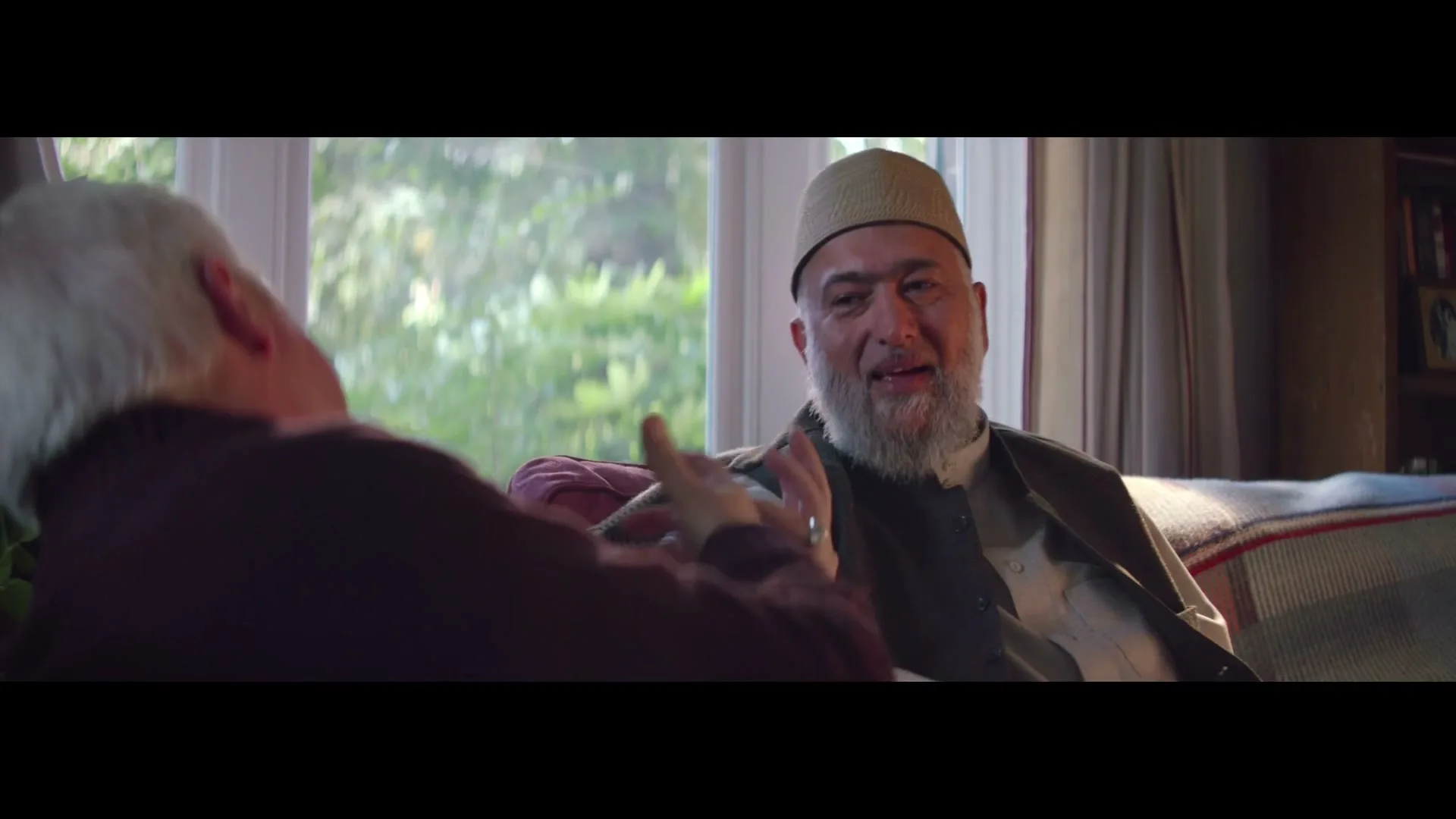 Two older men, one with a beard and a cap, sit on a couch and talk, gesturing with their hands in a cozy living room.