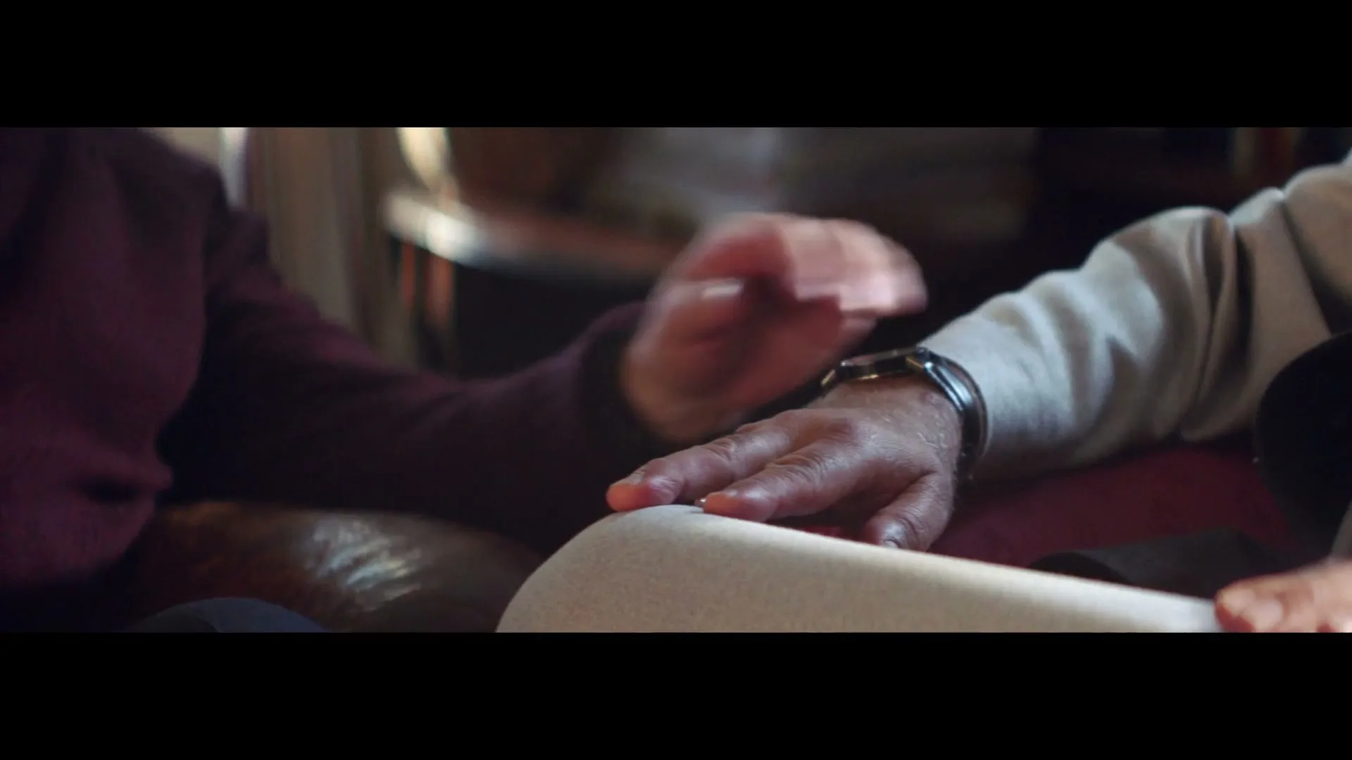 A close-up shot shows two people's hands, one wearing a watch, gently touching each other in a warm, intimate gesture indoors.