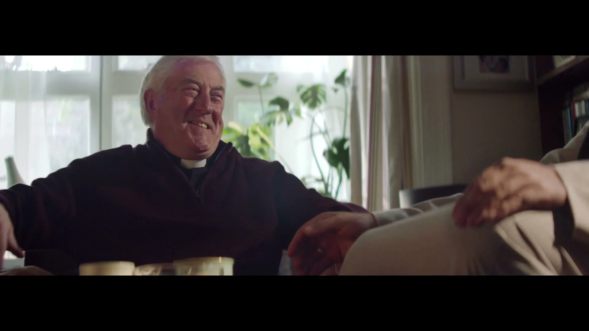 A senior male priest in a dark sweater laughs heartily while sitting on a couch next to another person, in a cozy living room.