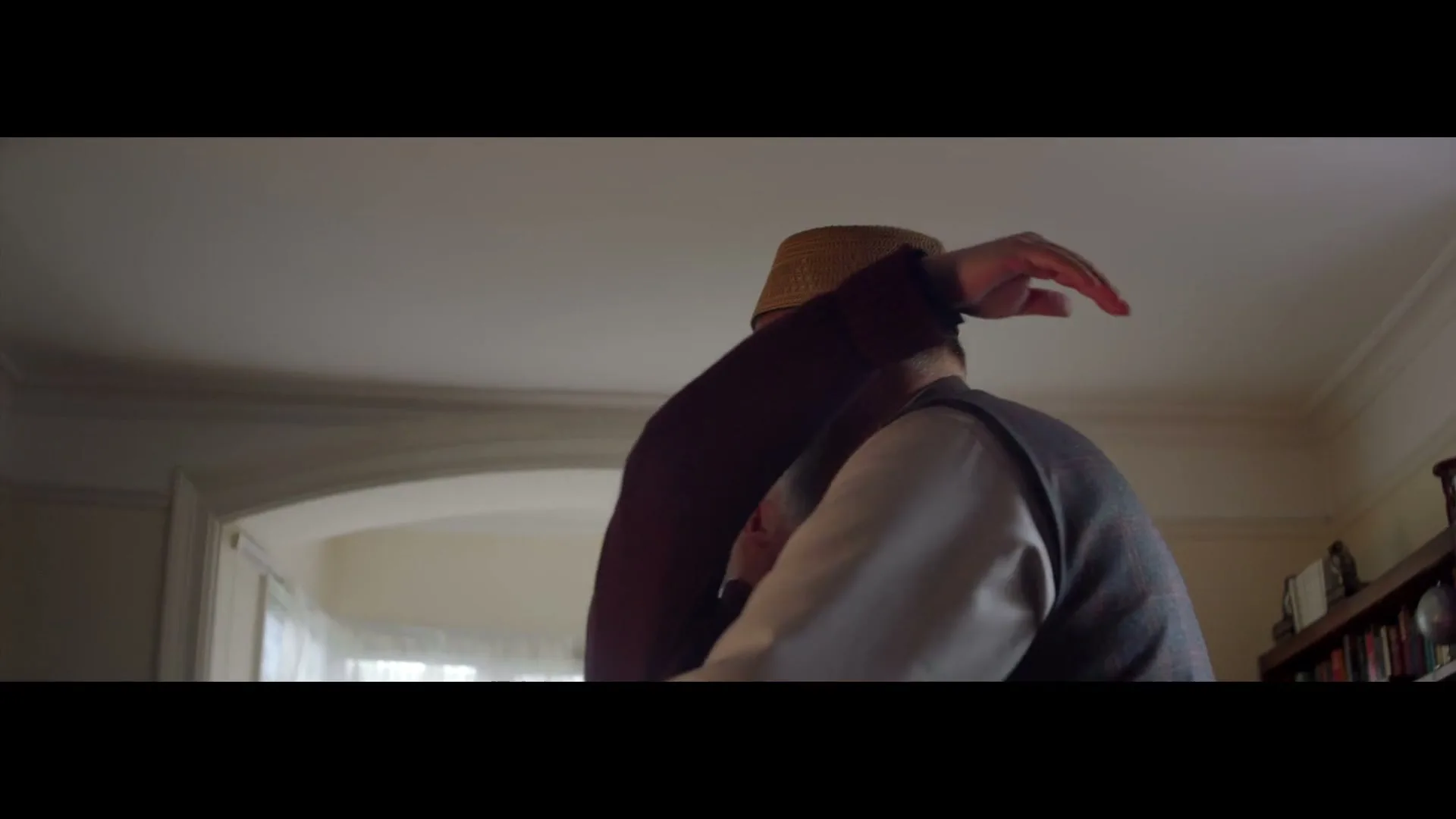 Two elderly men, one in a religious collar and the other in a kufi, embrace warmly in a sunlit living room.