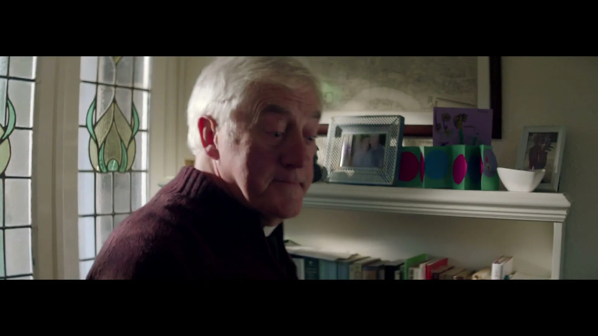 An elderly white man with white hair and a purple sweater turns to face the camera, then looks out a stained glass window in a cozy living room.
