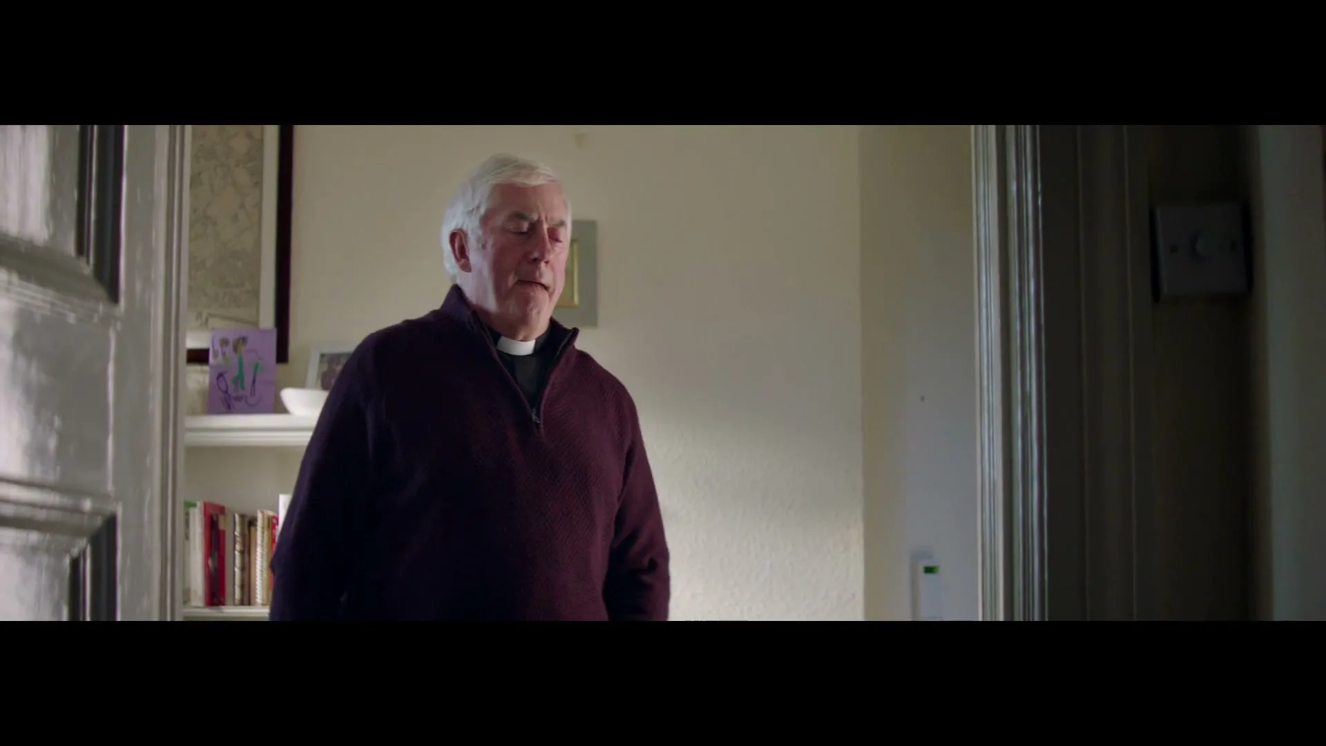 An older white man with white hair and a clerical collar walks into a room, then stops to look at his phone.