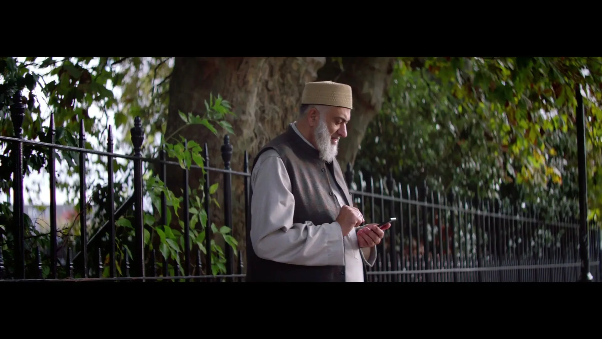 An older man with a white beard and a cap stands next to a black fence, looking down and interacting with his smartphone in a park.