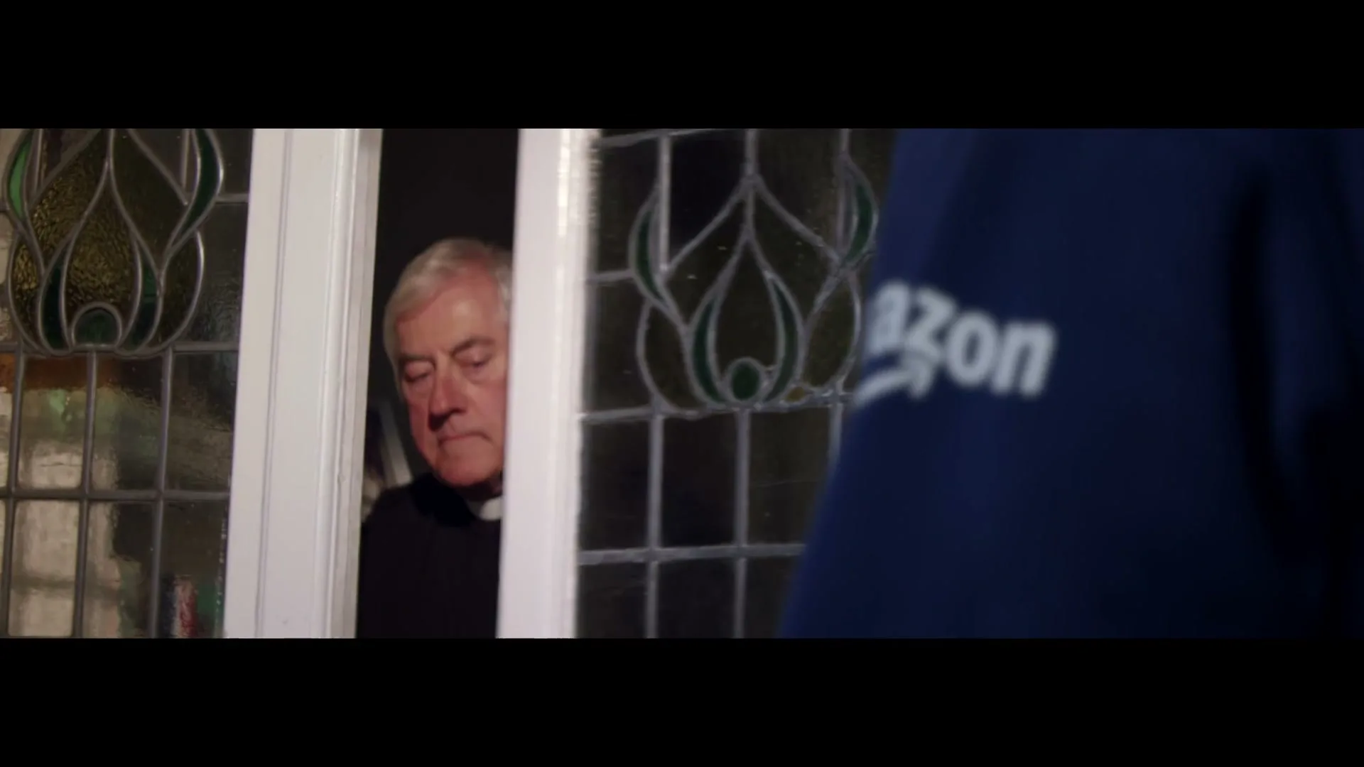 An Amazon delivery driver in a blue uniform hands a package to an older man in a black shirt at the front door of a house.