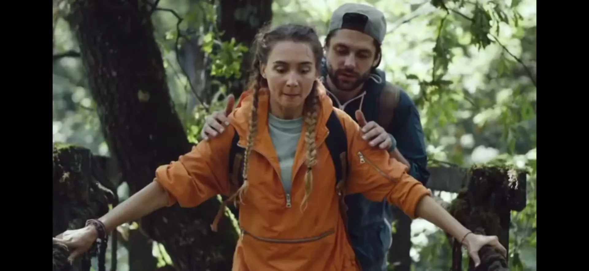 A man helps a woman with braided hair in an orange jacket cross a wooden bridge in a lush green forest.