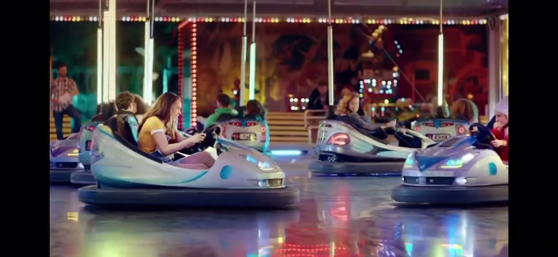 A young woman in a yellow shirt drives a bumper car at a brightly lit amusement park, surrounded by other people in bumper cars.