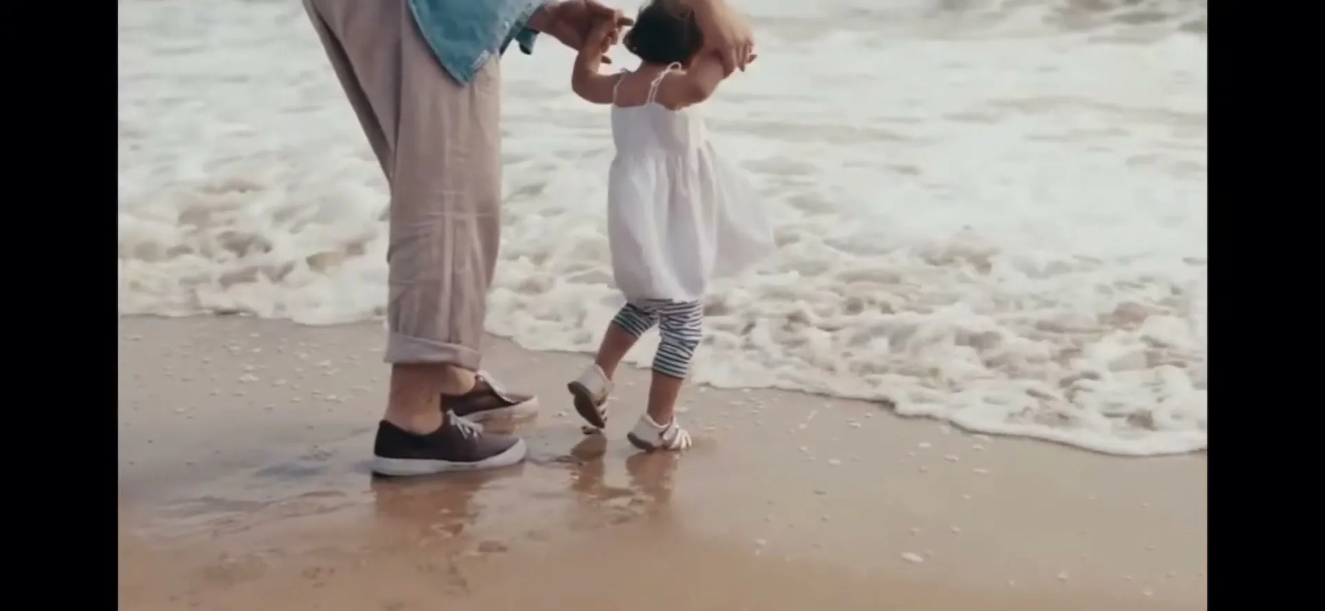 A father holds his young daughter's hands as she takes steps on a sandy beach with ocean waves washing over their feet.