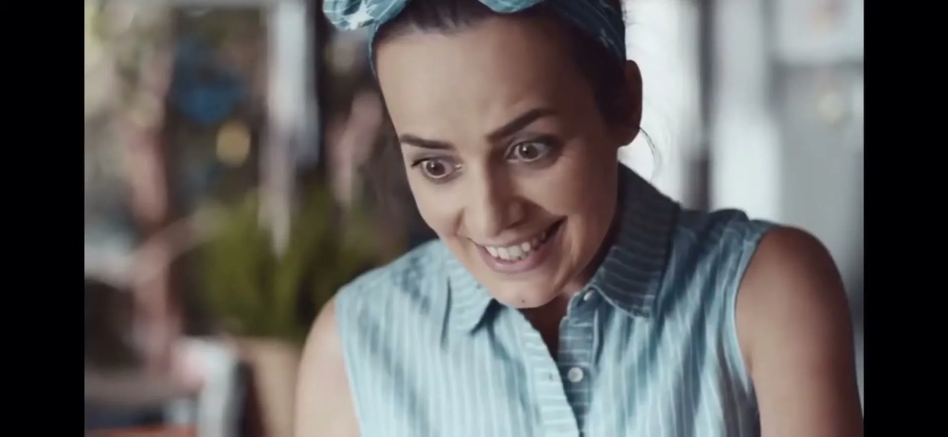 A woman with a blue headscarf and striped shirt looks down with a surprised, happy expression in an interior setting.