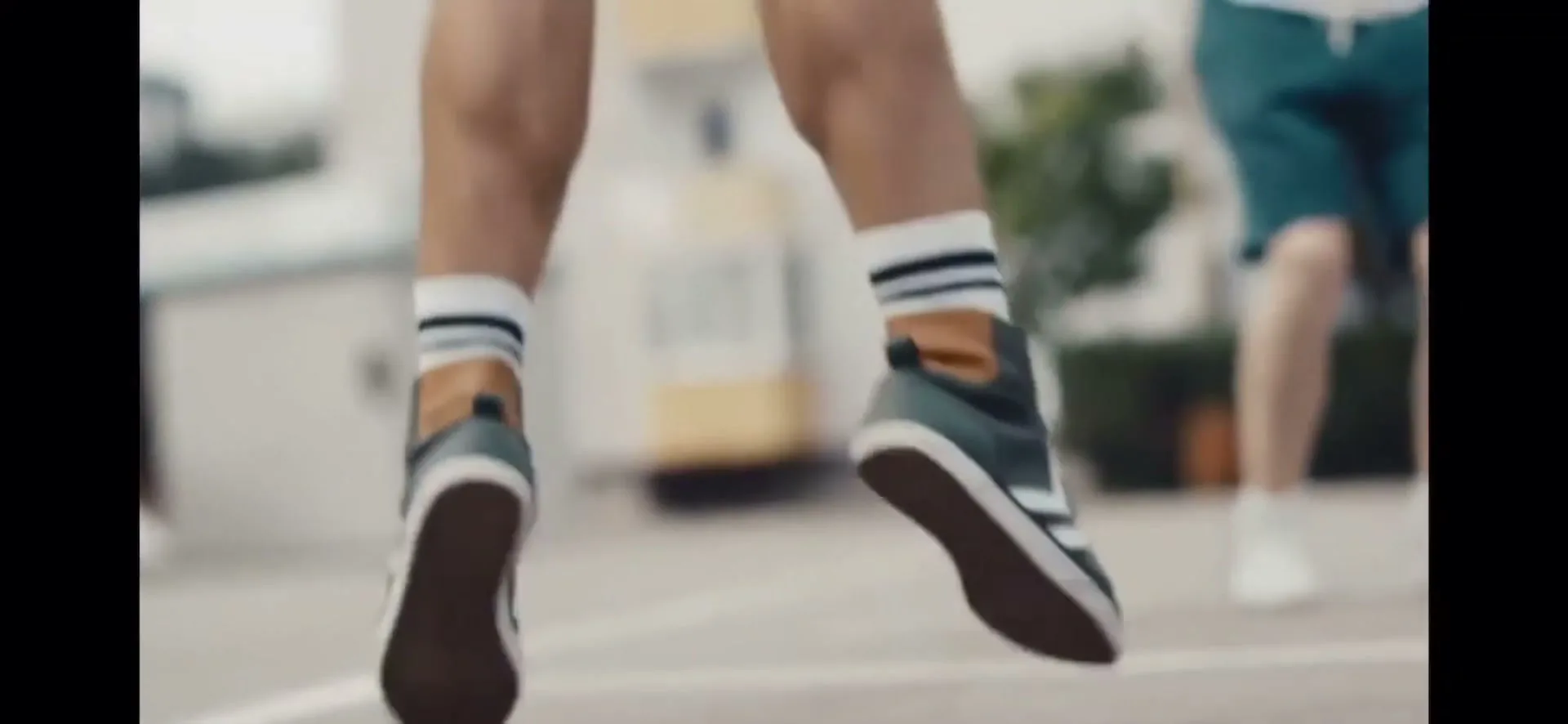 A person in shorts and sneakers jumps in the air, seen from behind, with another person partially visible in the background.