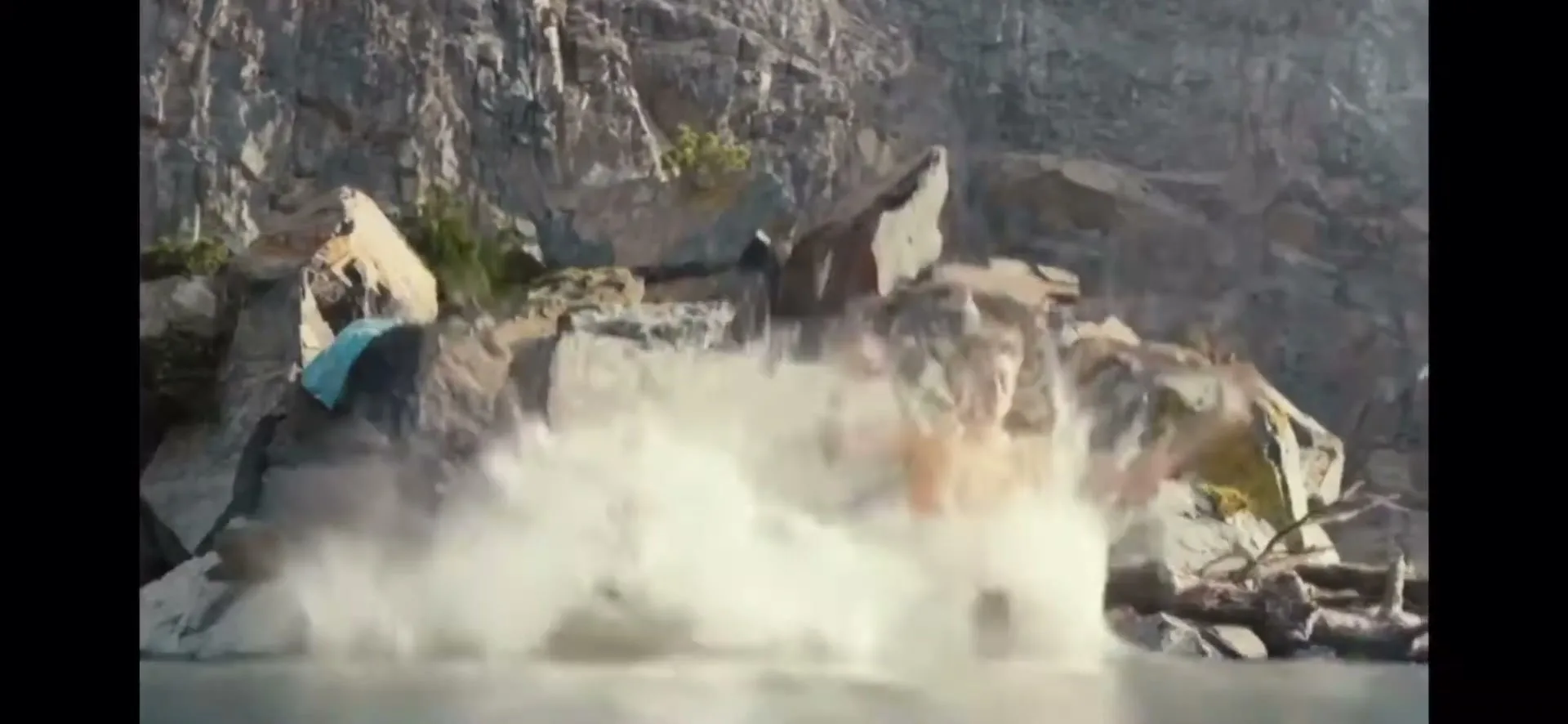 Two young men in swim trunks jump off a rocky cliff into a lake, creating a large splash in a sunny outdoor setting.