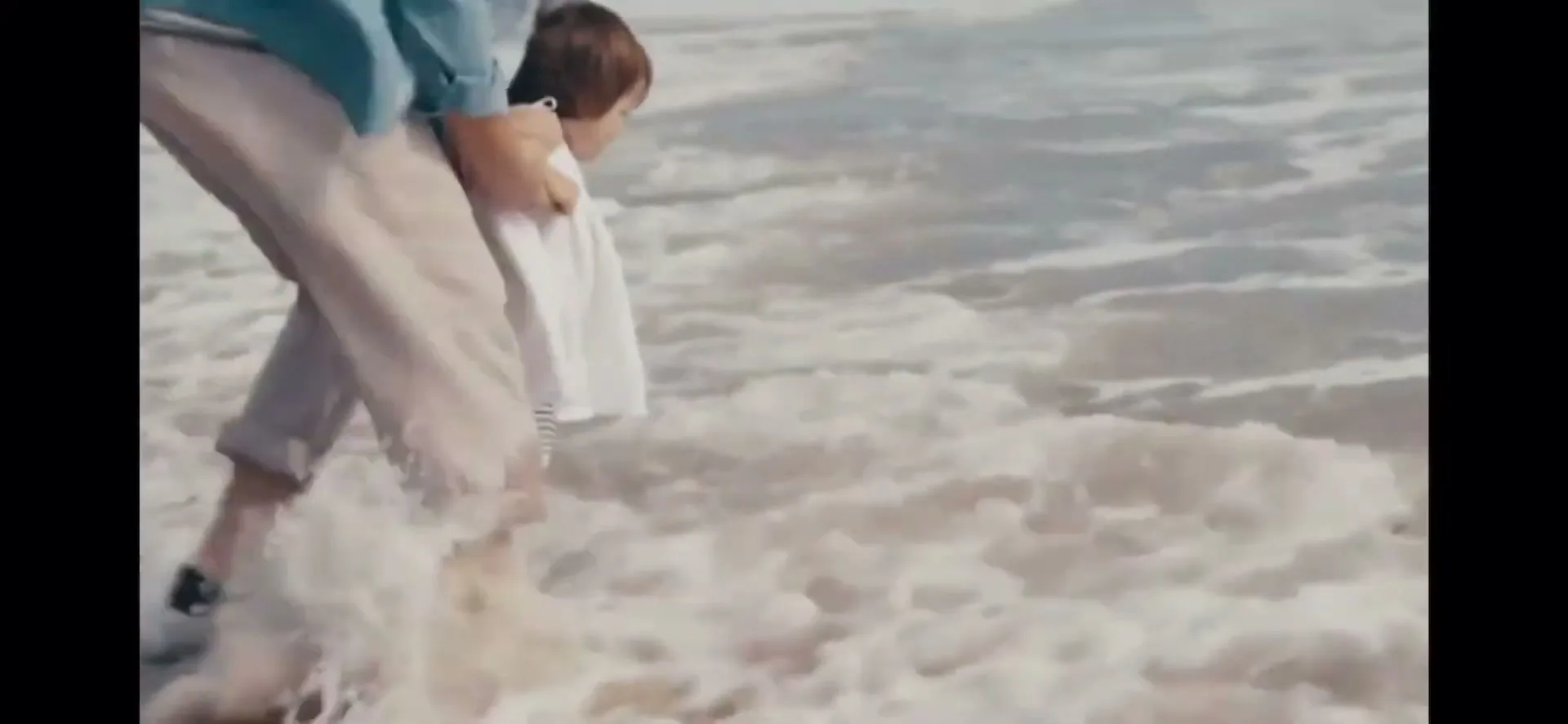 A person in a blue shirt and khaki pants holds a small child in a white top as they stand in the foamy ocean water at a beach.