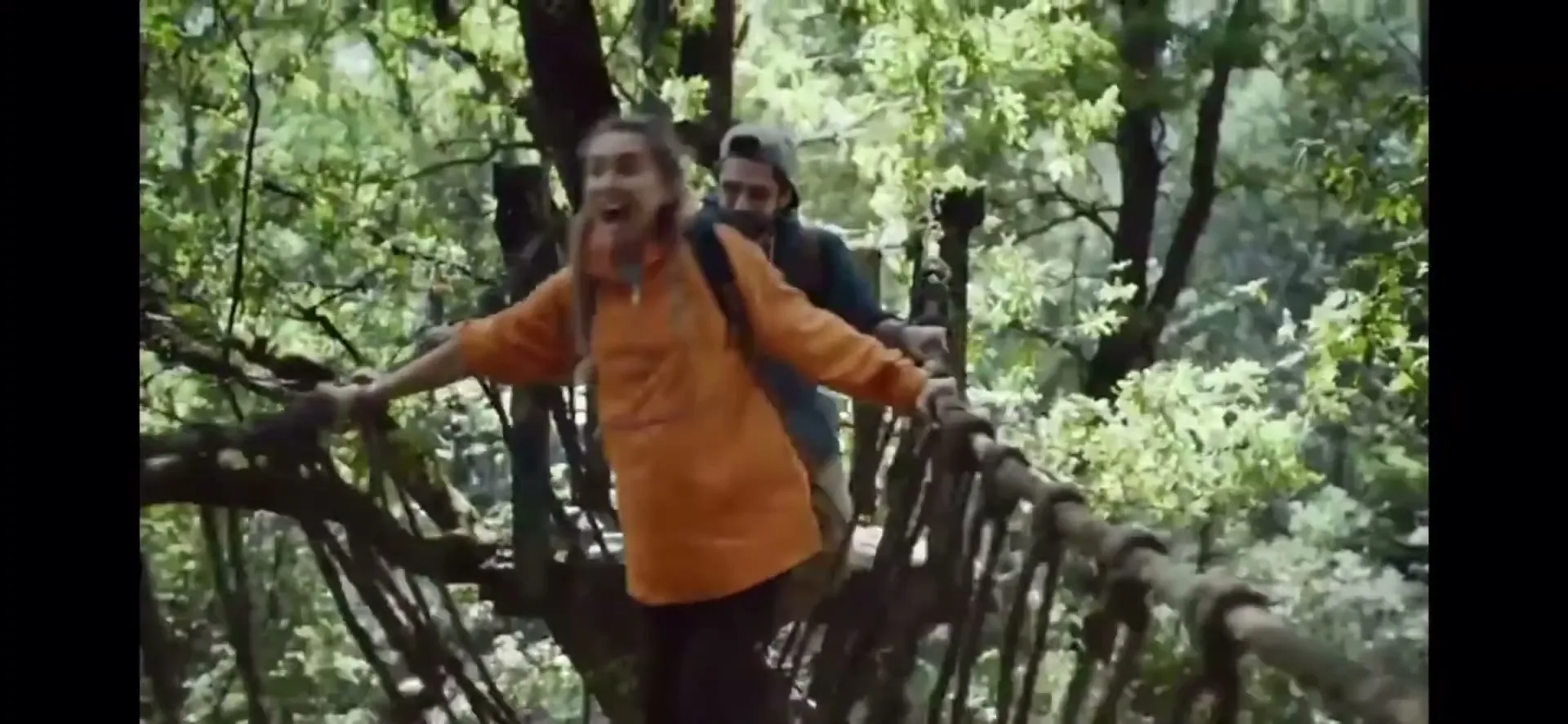 A young woman in an orange jacket and backpack crosses a rope bridge in a lush green forest, with a man following behind her.