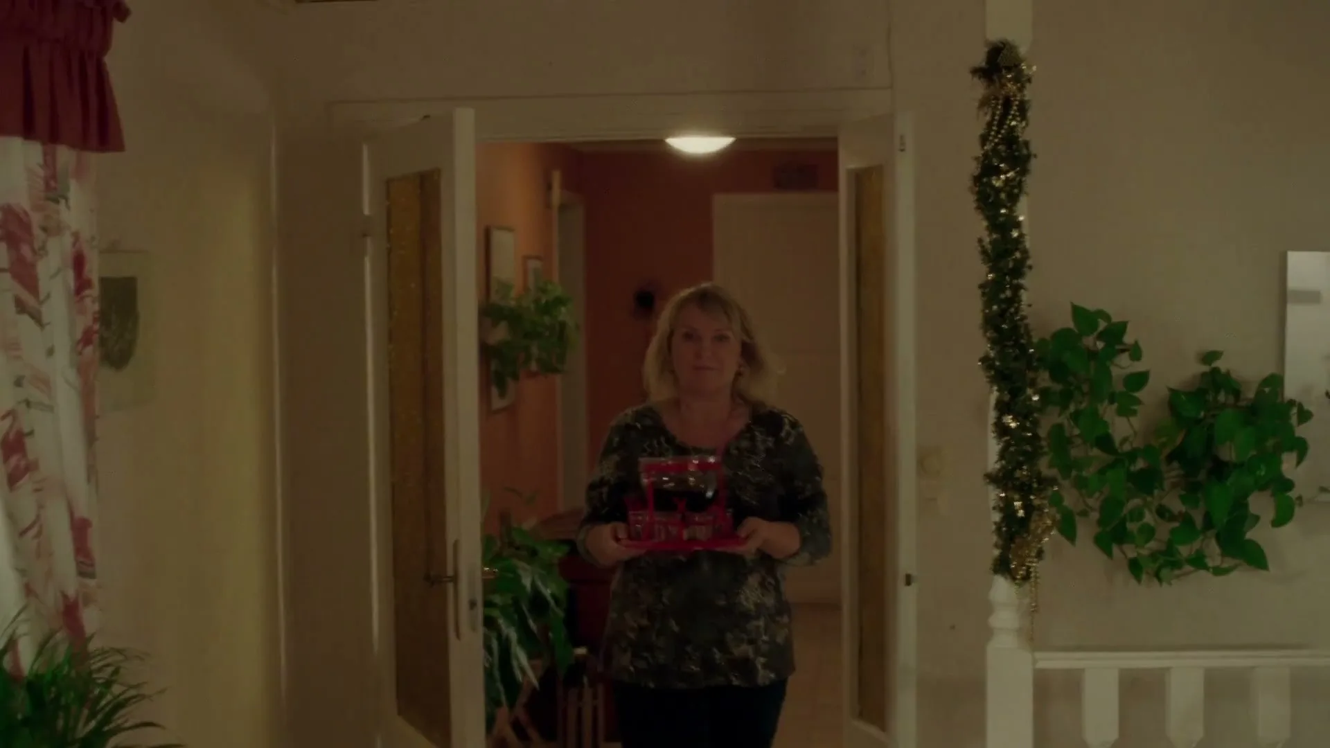 A middle-aged woman in a patterned top carries a red tray with drinks, walking through a doorway in a home decorated for Christmas.