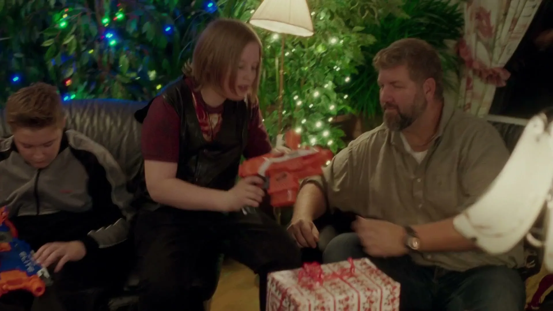 A young boy with long hair in a black vest and red shirt shows a toy gun to a bearded man, while another boy looks at his own toy gun, in a living room decorated for Christmas.