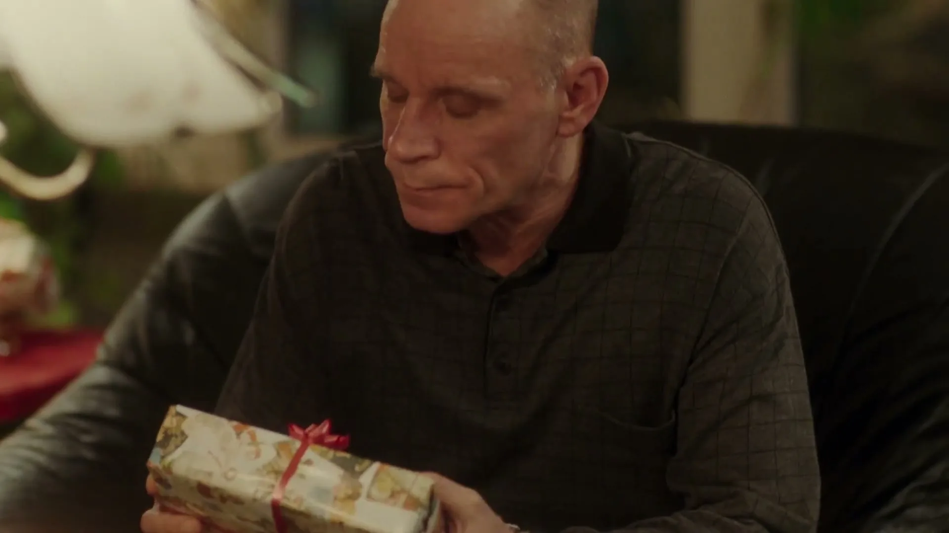 A middle-aged man with a shaved head sits on a black leather couch, holding a wrapped Christmas gift, looking around with a slightly confused expression in a cozy living room.