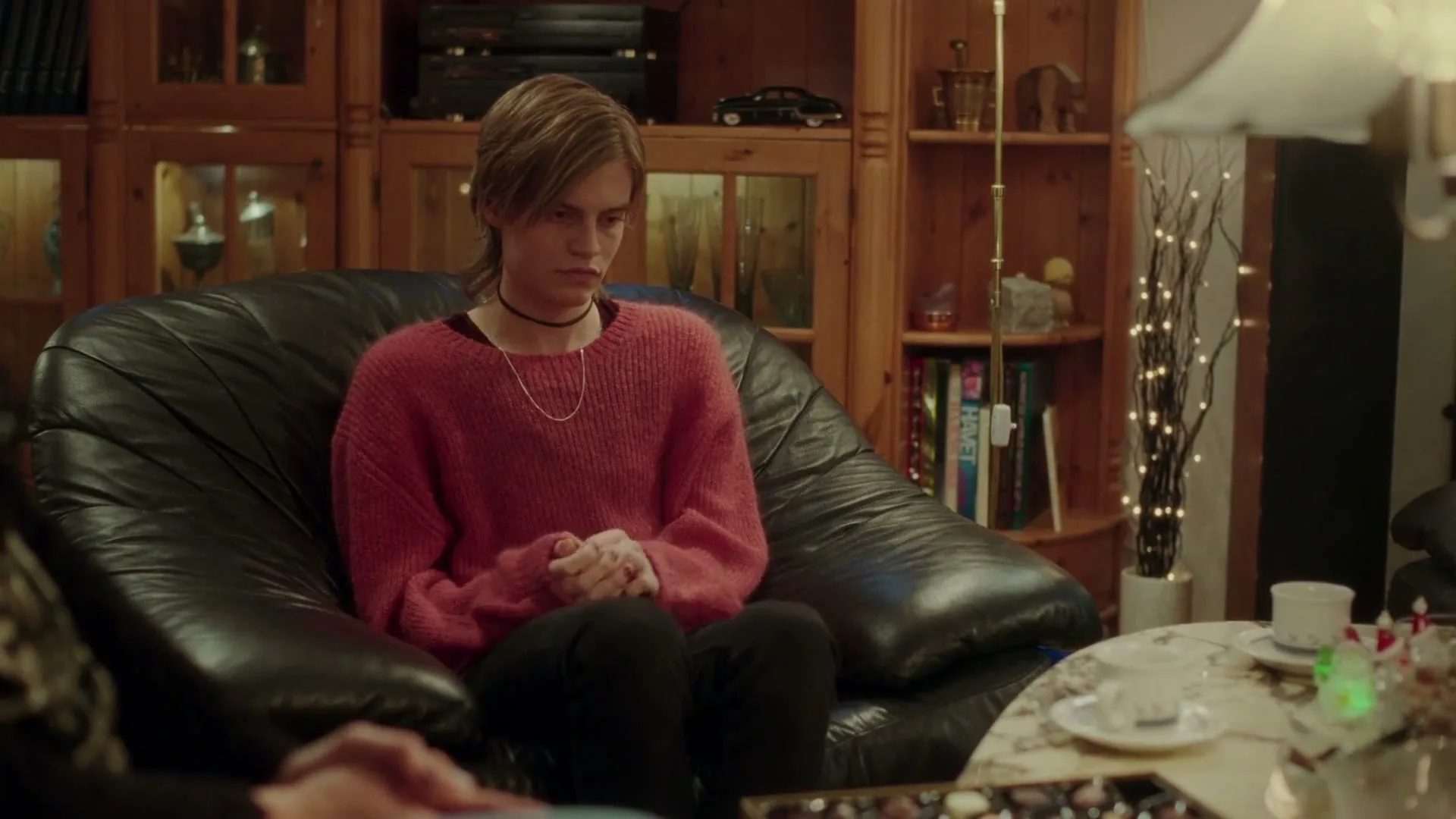 A teen with long hair and a pink sweater sits on a black leather armchair, looking down at their hands in a cozy living room.