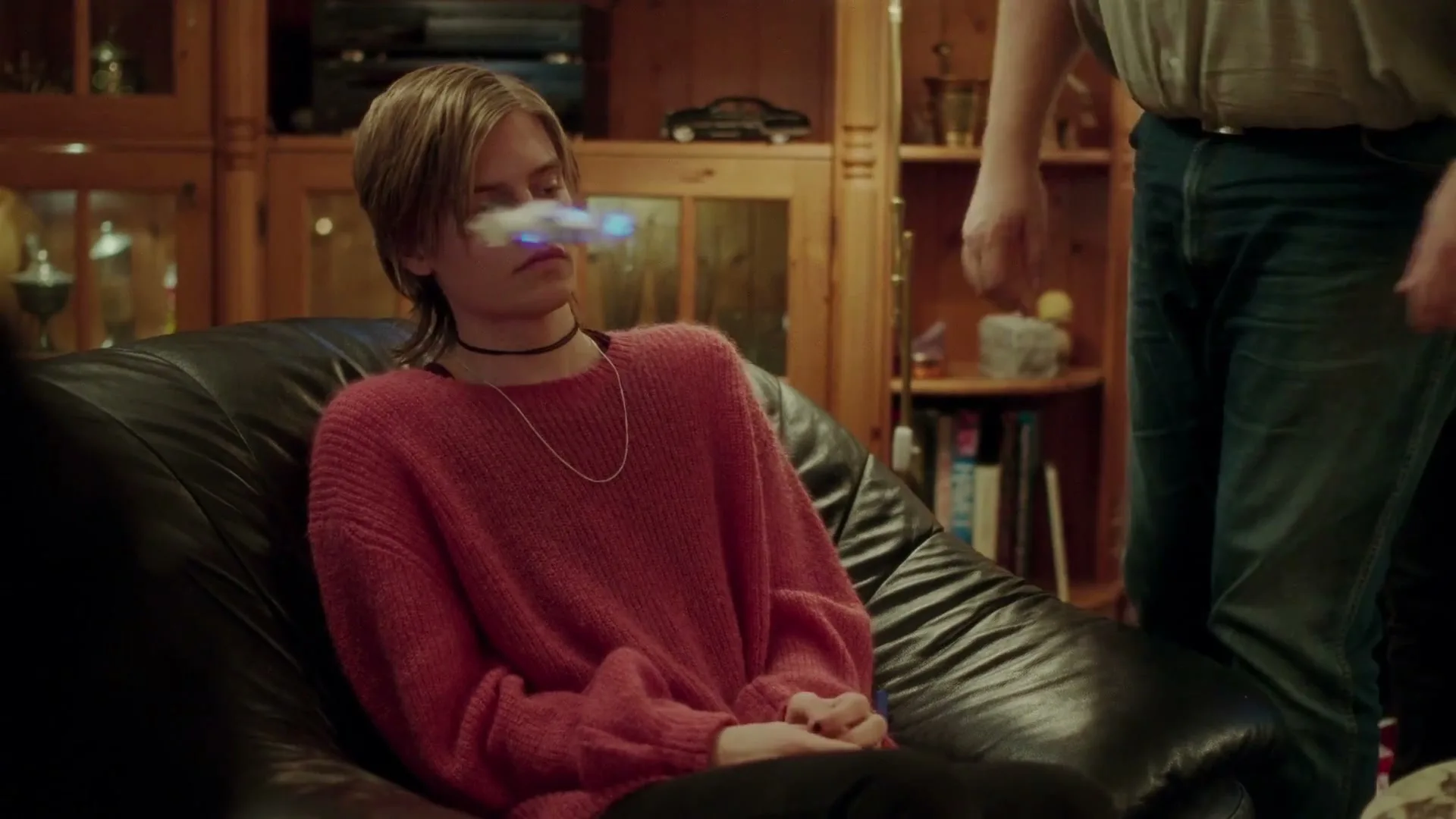 A teen with short hair and a pink sweater sits on a black leather chair, looking bored as a small drone flies past their face in a cozy living room.