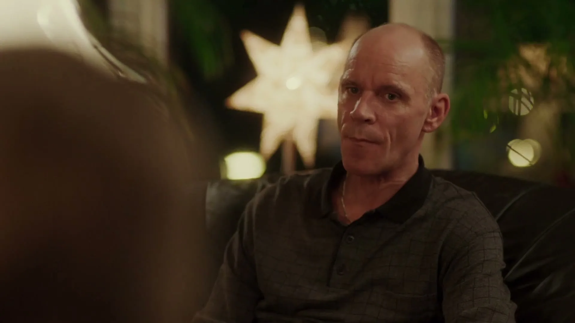 A bald man in his 50s sits on a couch, looking off-camera with a neutral expression in a dimly lit living room.