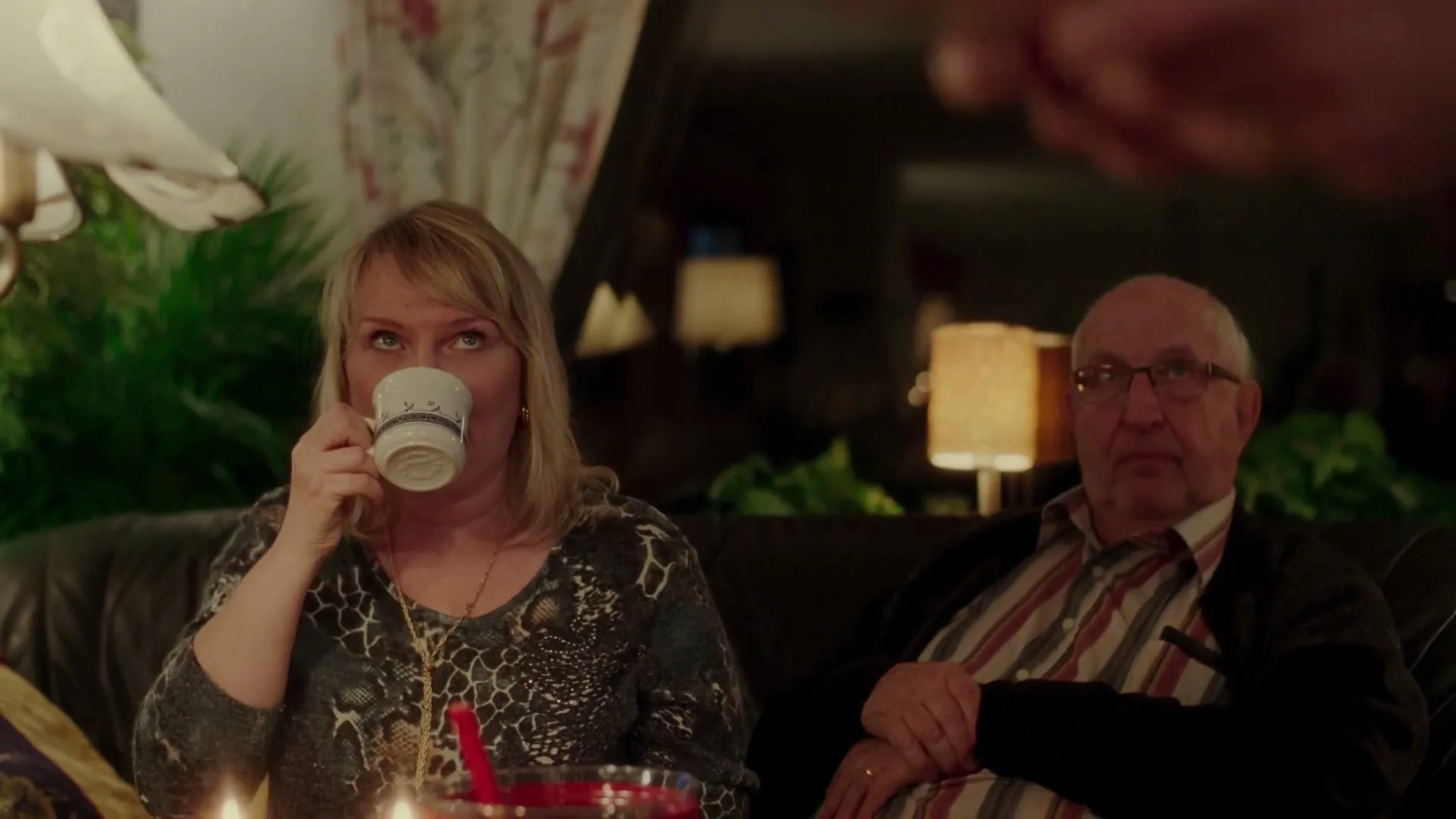 A blonde woman sips from a mug while sitting on a couch next to an older man, both looking off-camera in a dimly lit living room.