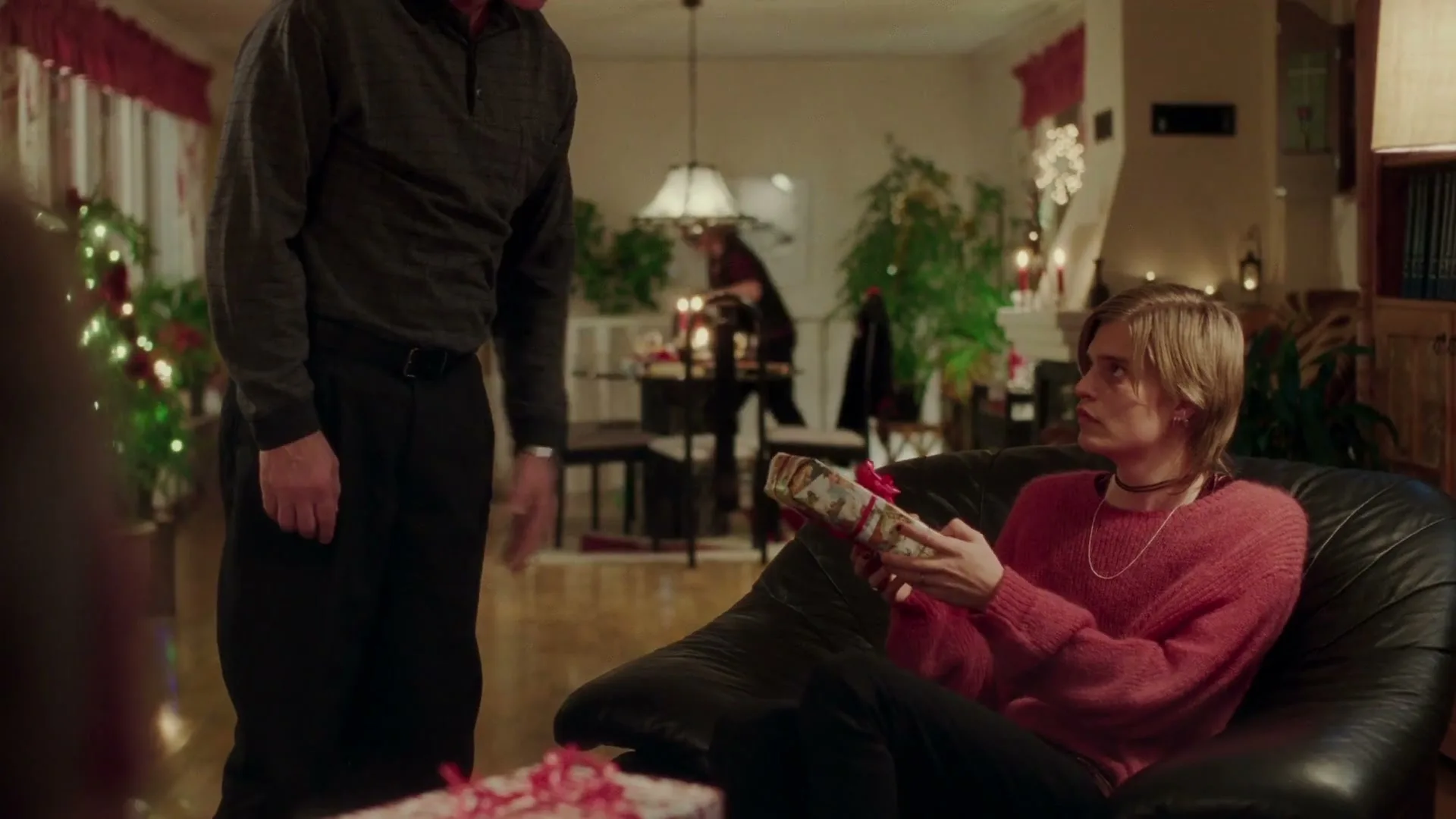 A man in a dark shirt hands a wrapped gift to a young person with blonde hair in a red sweater, sitting on a black couch in a festive living room.