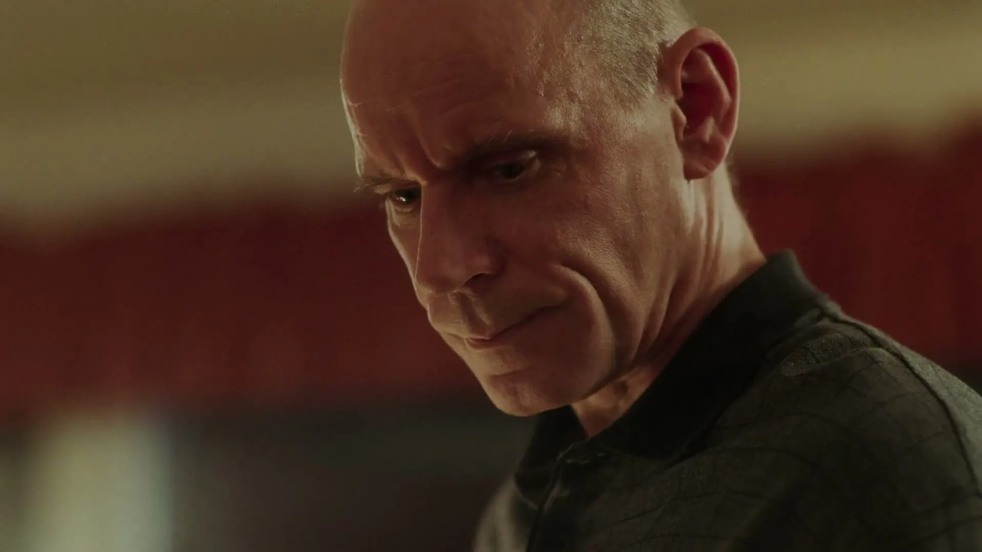 A bald man in his 50s or 60s, wearing a dark polo shirt, looks down with a concerned expression in an interior setting.