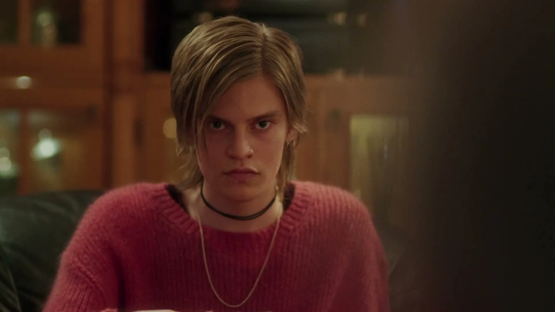 A teen with blonde hair and a pink sweater looks up with a serious expression in a dimly lit living room.