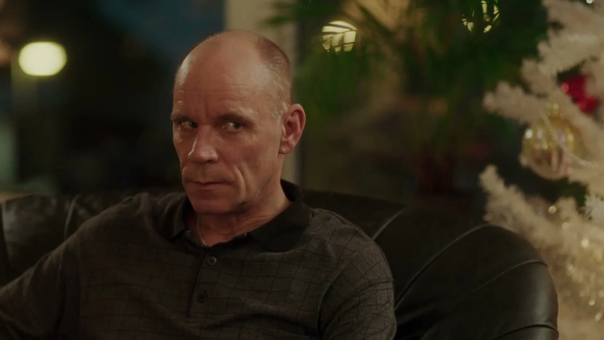 A bald man in his 50s sits on a black leather couch, looking concerned, next to a white Christmas tree in an interior setting.